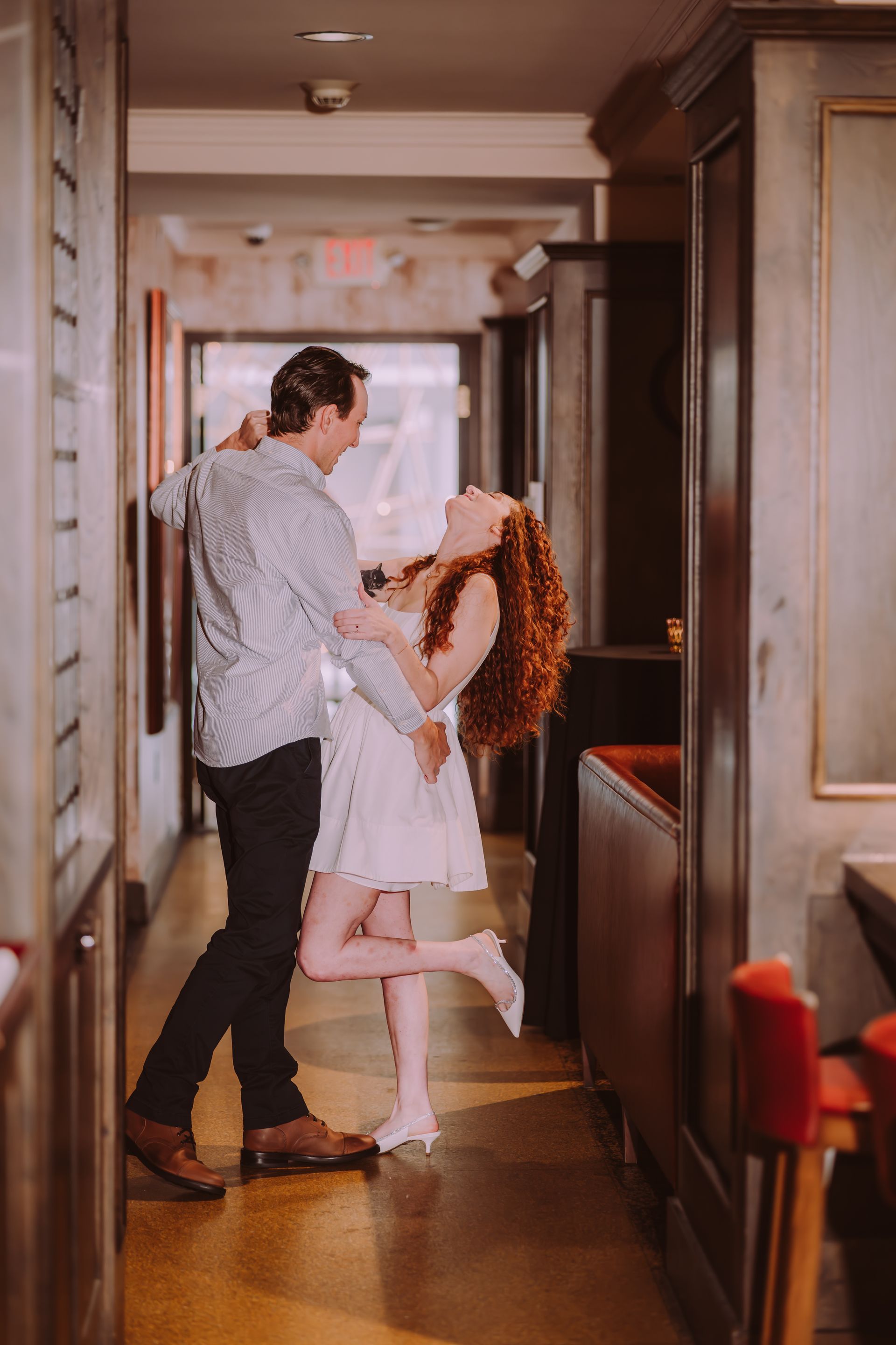 Couple dancing joyfully in a restaurant hallway, woman in white dress, man in blue shirt, reddish-brown hair.