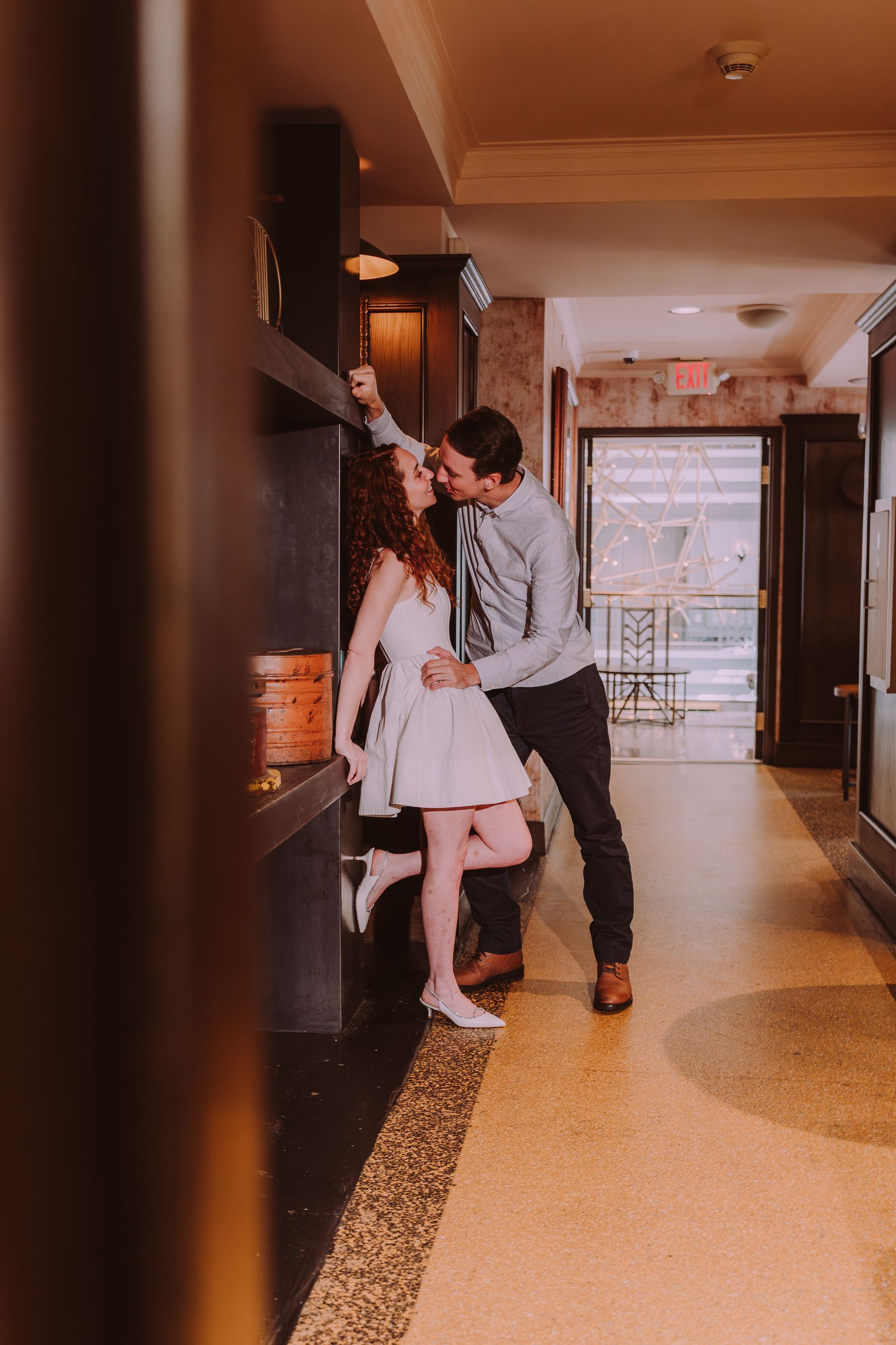 Couple embraces in a hallway; woman in white dress, man in blue shirt, near a dark wall.