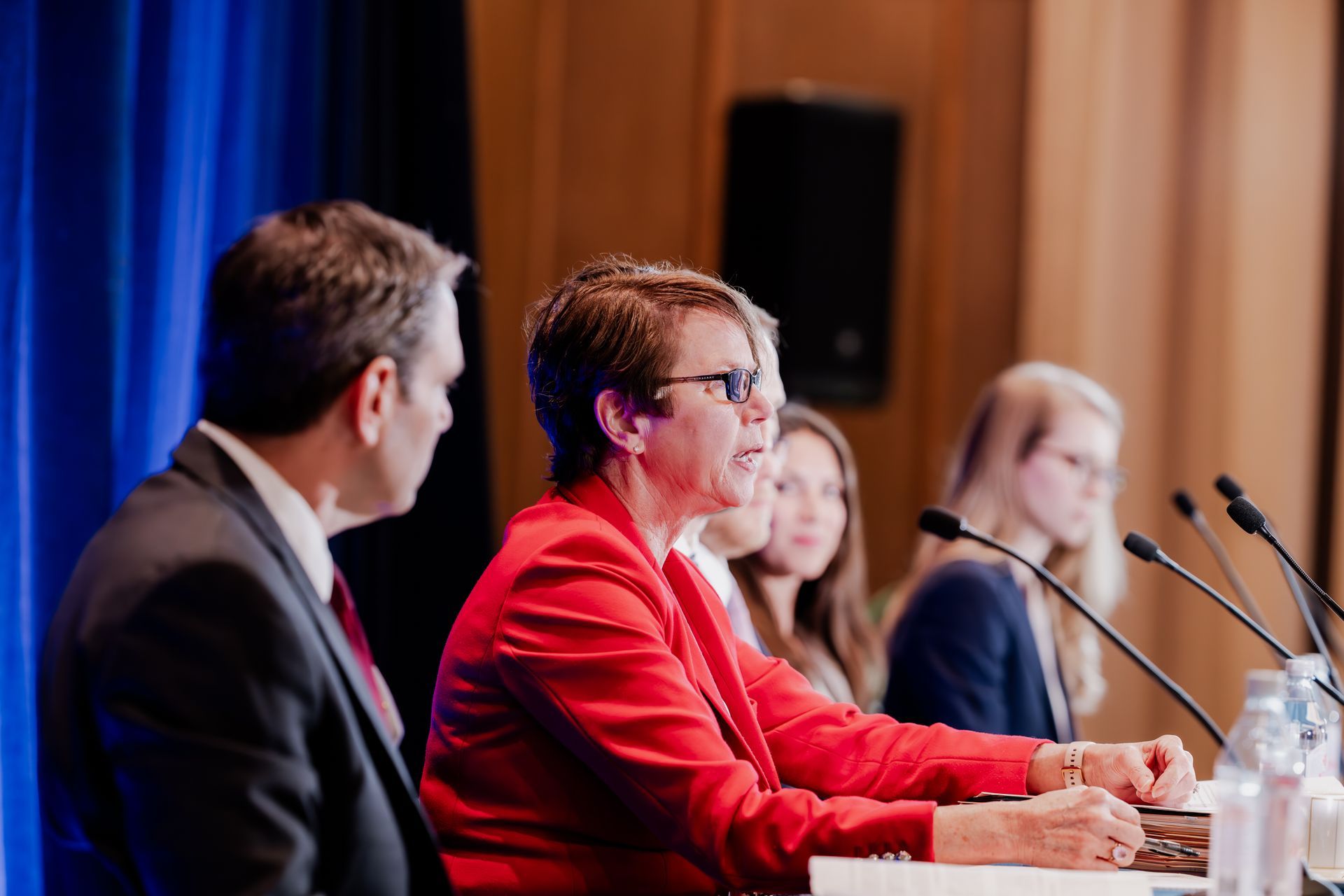 Panel of people at a table, microphones, speaker in red blazer.