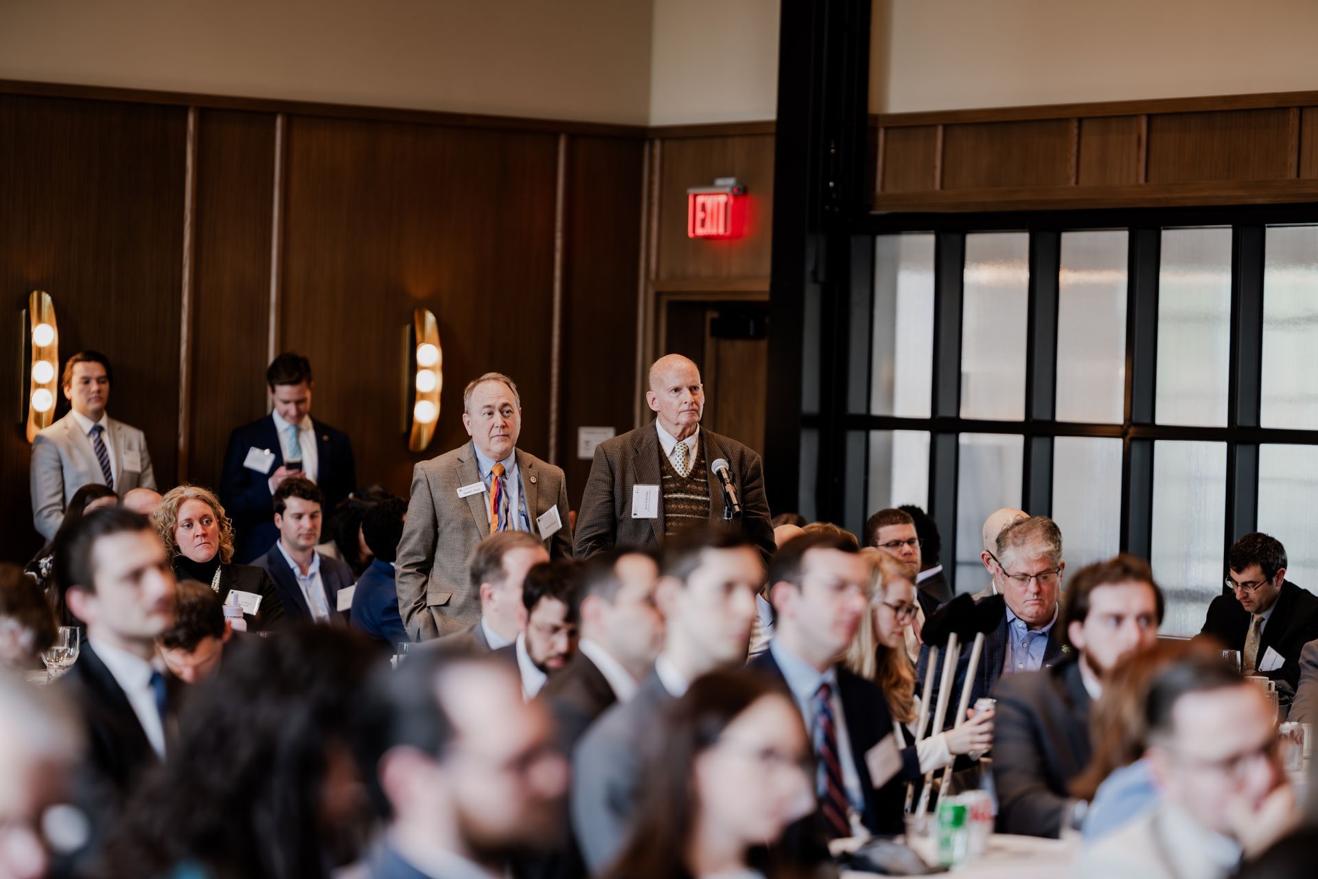 An audience listens to speakers at an indoor conference held in a room with wood-paneled walls and an exit sign.