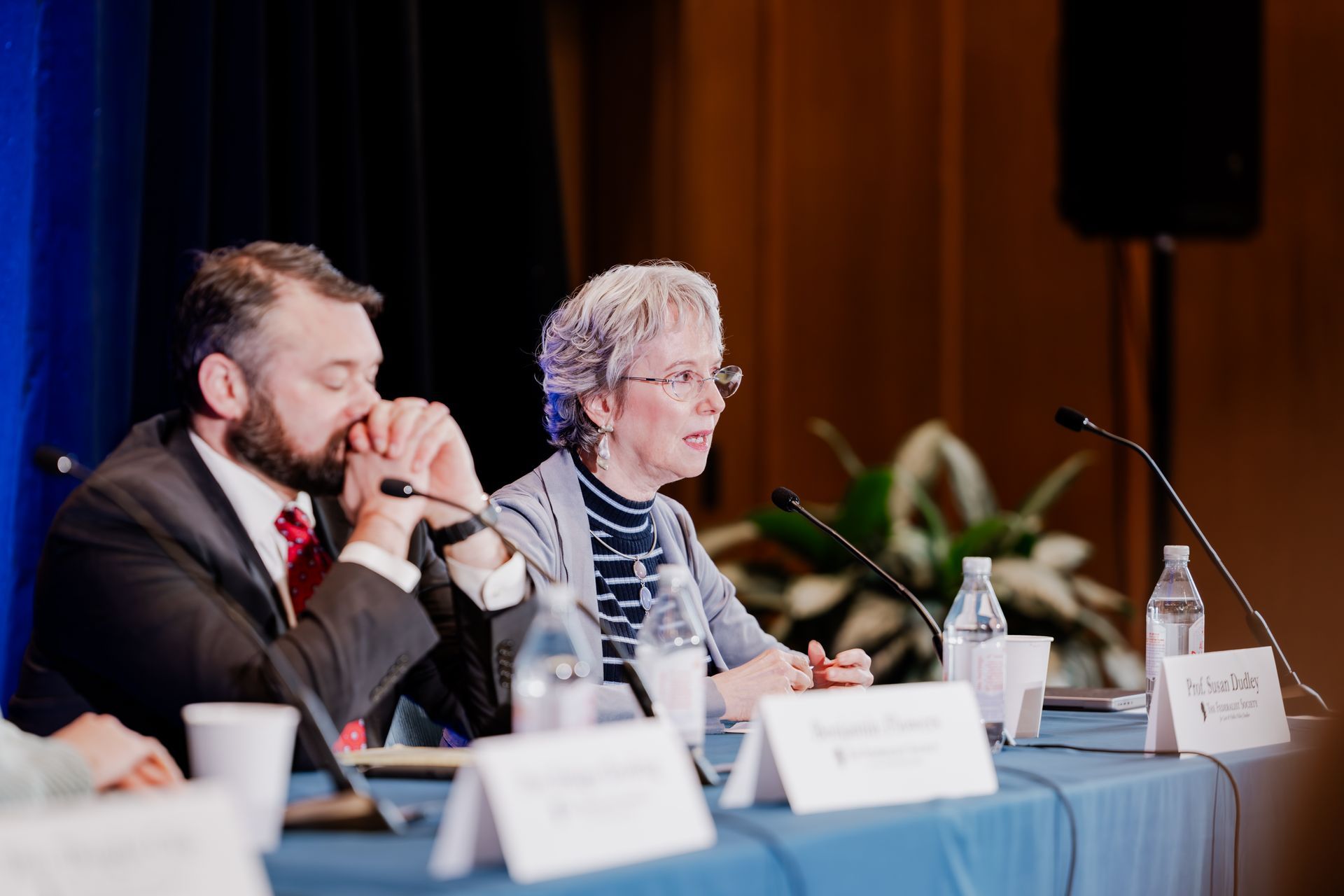 Two people sit at a table with microphones and nameplates during a panel discussion in a conference setting.