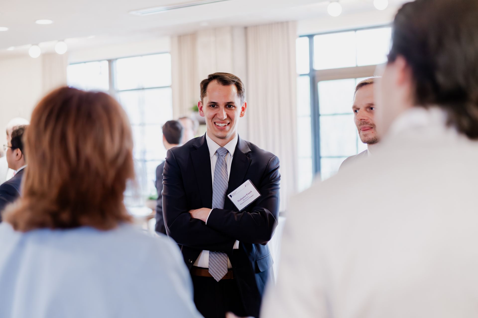 Man in suit smiles, arms crossed, conversing with others at an event. Light-filled room.