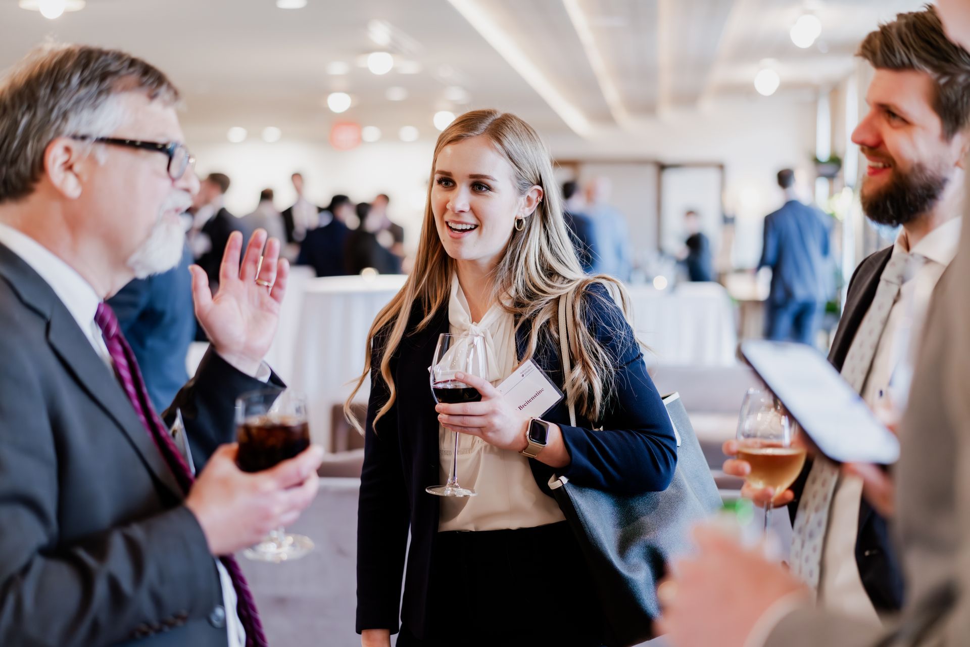 Three people in professional attire chat and hold drinks at a bright indoor business networking event.