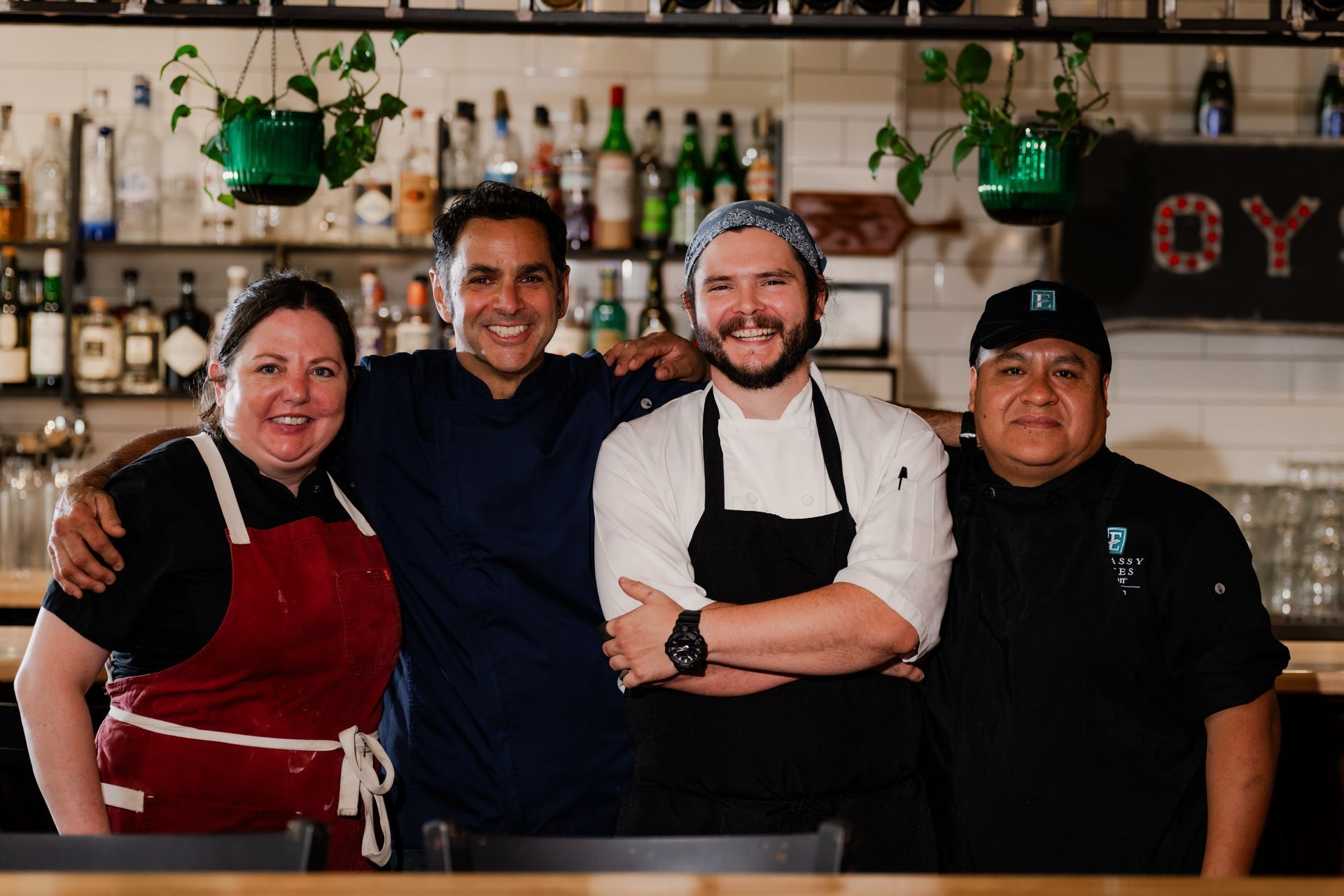 Four restaurant staff smiling, arm-in-arm. Behind them, a bar with bottles and plants.