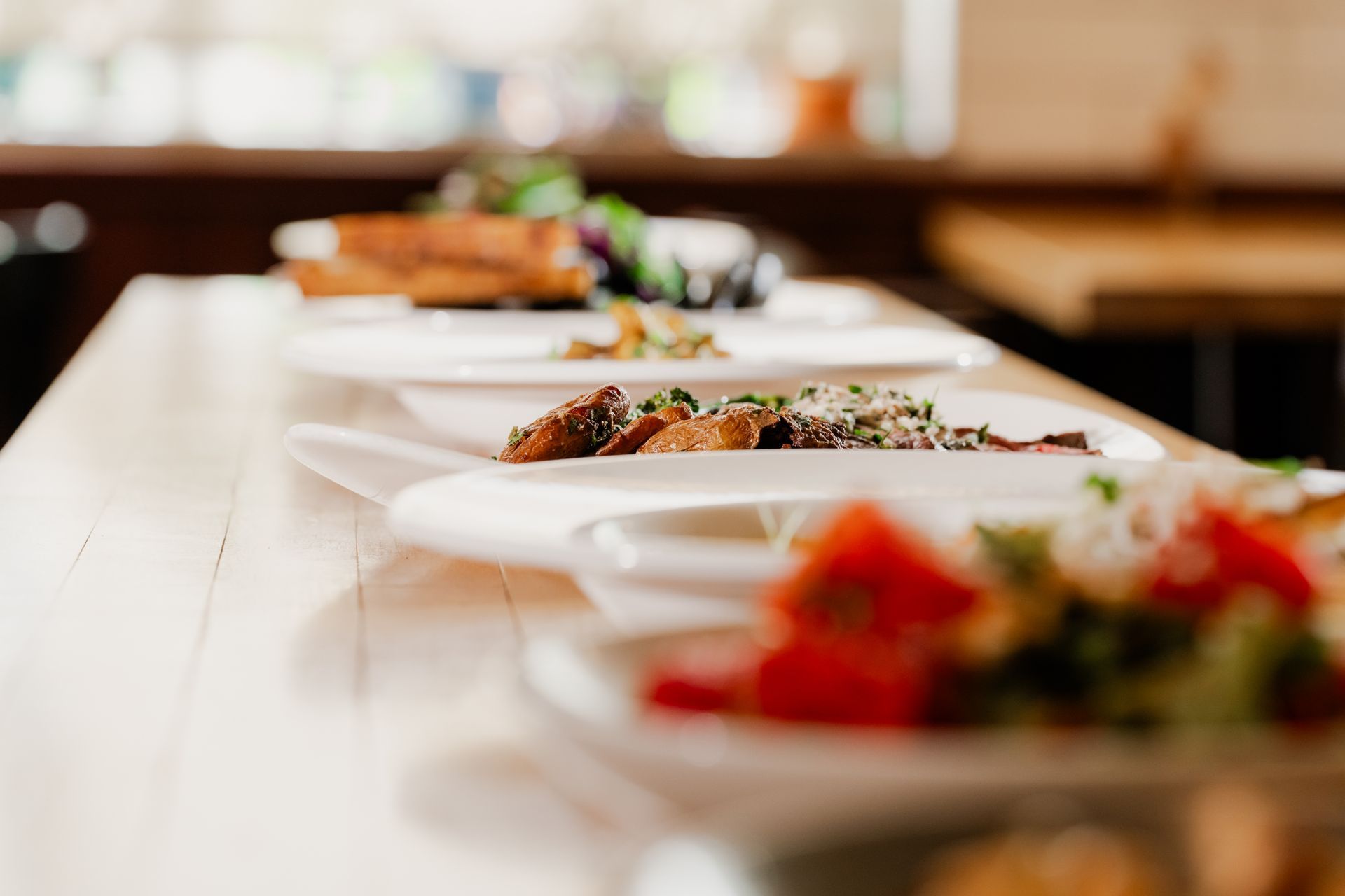 Plates of food lined up on a wooden surface, restaurant setting; tomatoes, grains, and meat visible.