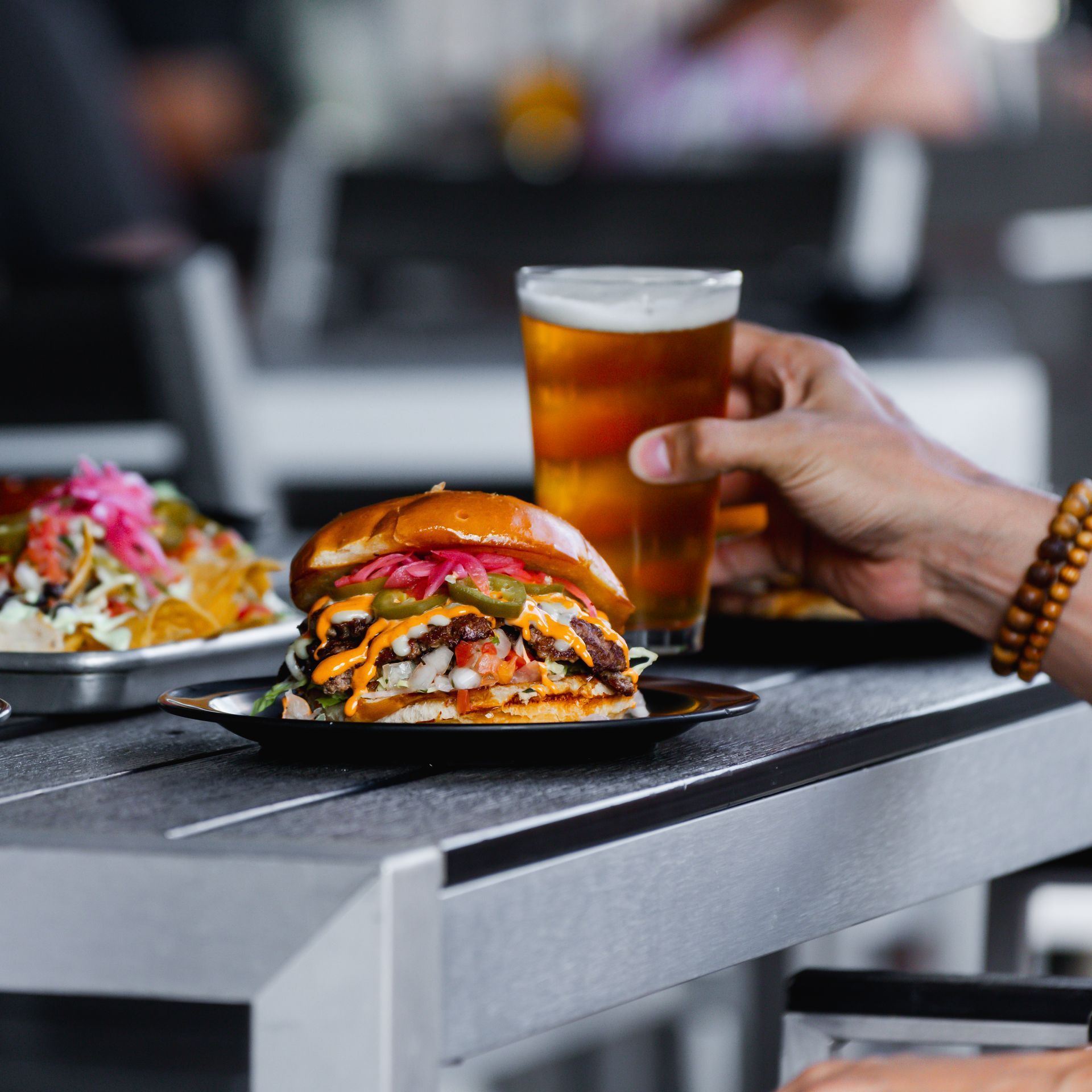 A hand holding beer next to a gourmet burger and plate of nachos on a table, outdoor setting.