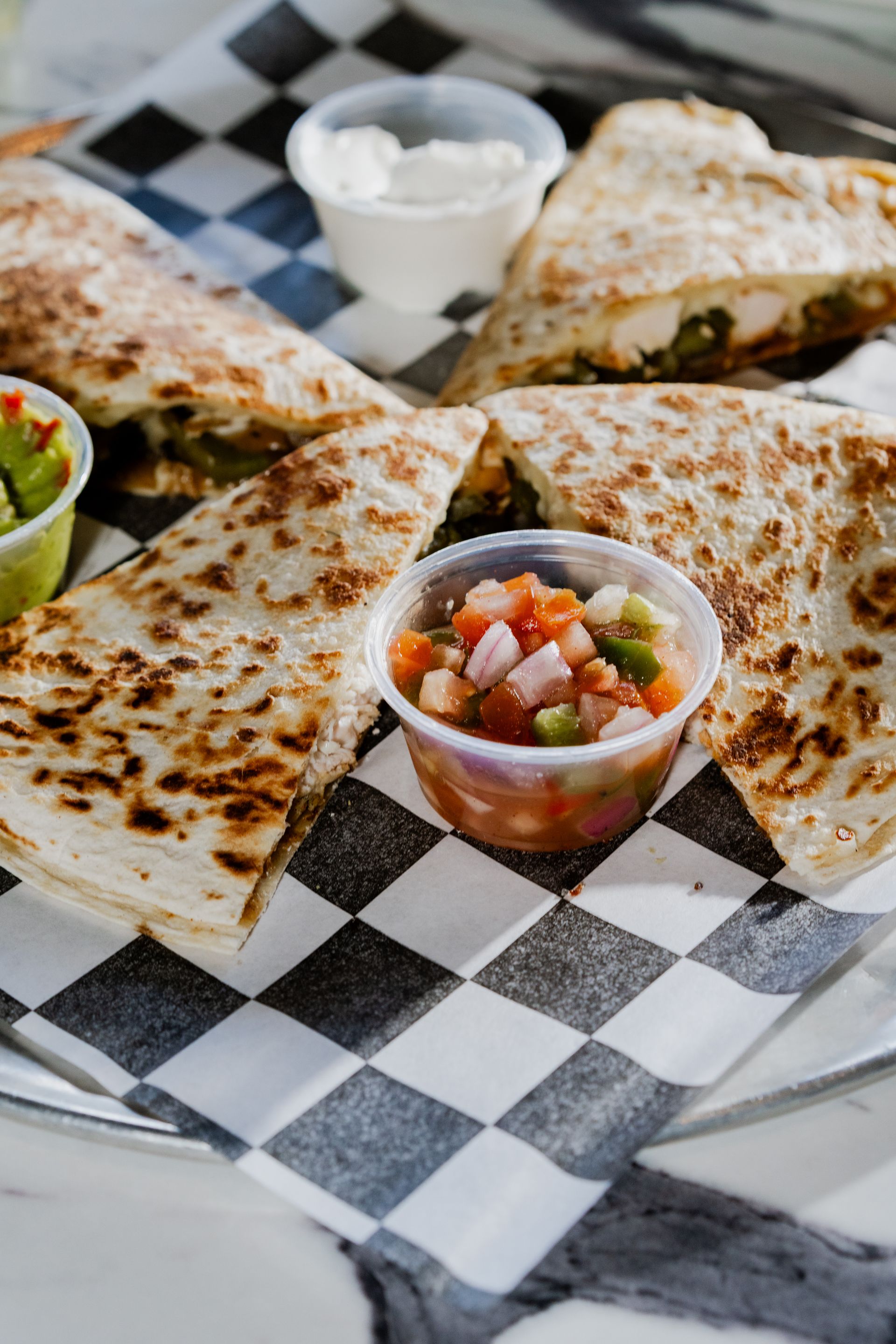 Quesadillas with salsa, sour cream, and guacamole, served on a checkerboard paper.