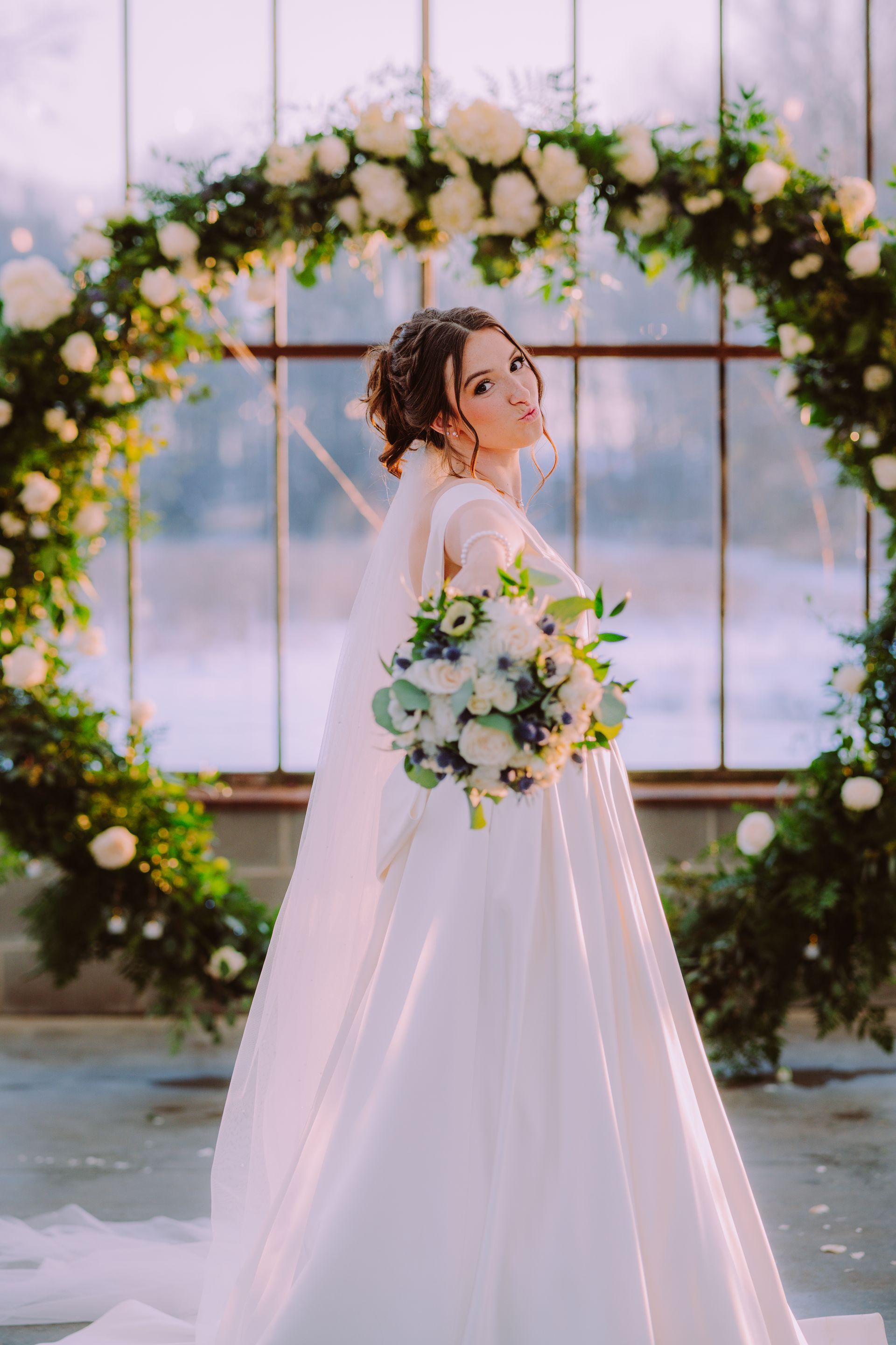 Bride in white dress holds bouquet, poses before floral arch in greenhouse.