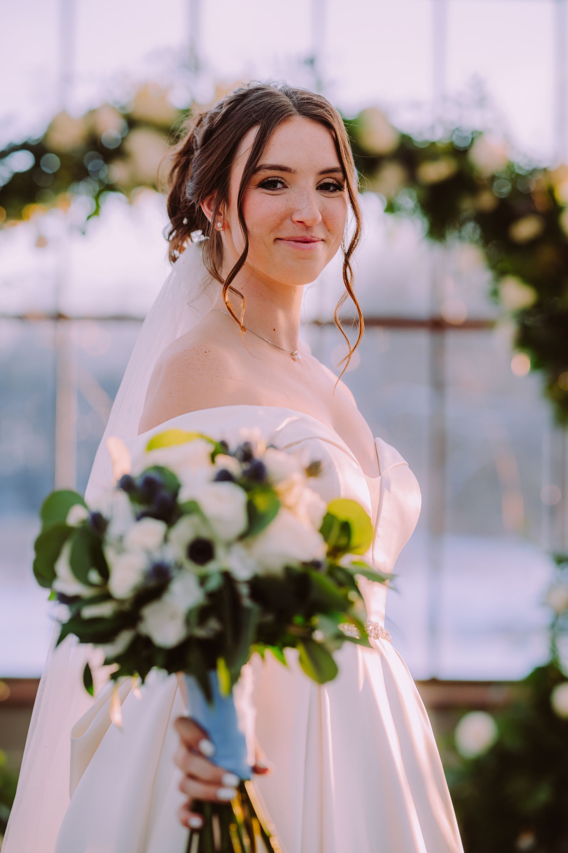 Bride holding a bouquet, wearing an off-the-shoulder gown, in front of a floral arch.