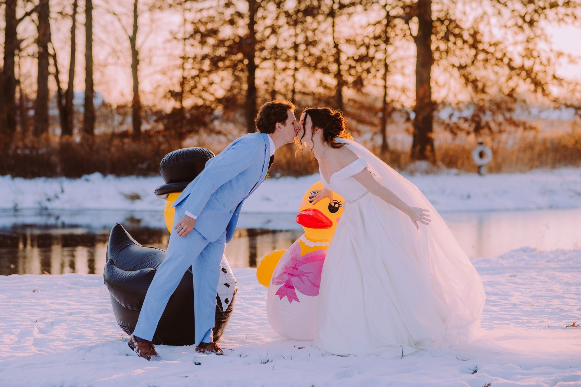 Bride and groom kissing outdoors in snow, next to inflatable penguin and duck. Sunset in background.