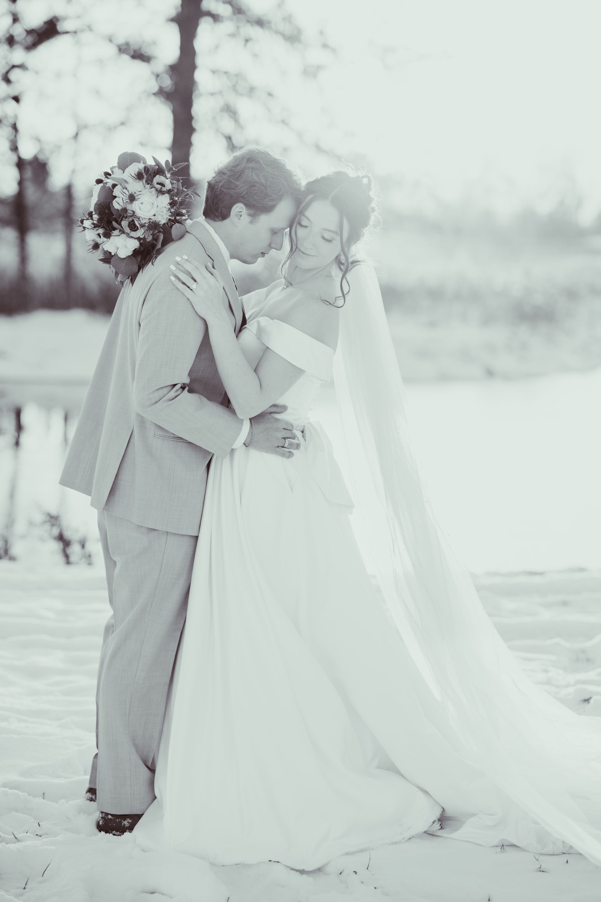 Bride and groom embrace on a beach. The bride wears a white dress and veil; the groom wears a suit.