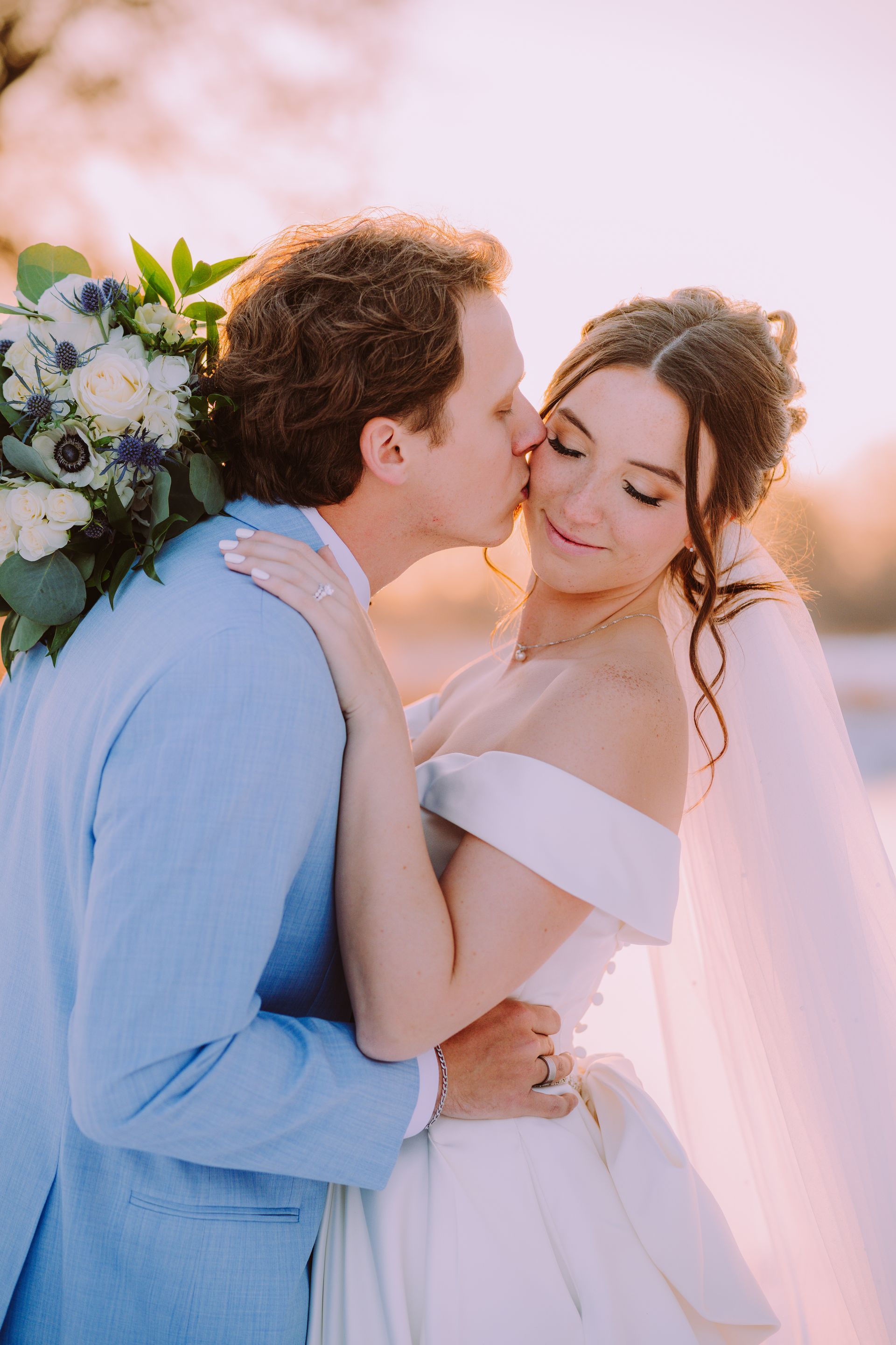 Groom kisses bride's cheek at sunset. She wears an off-shoulder gown, veil. He wears a blue suit, holds a bouquet.