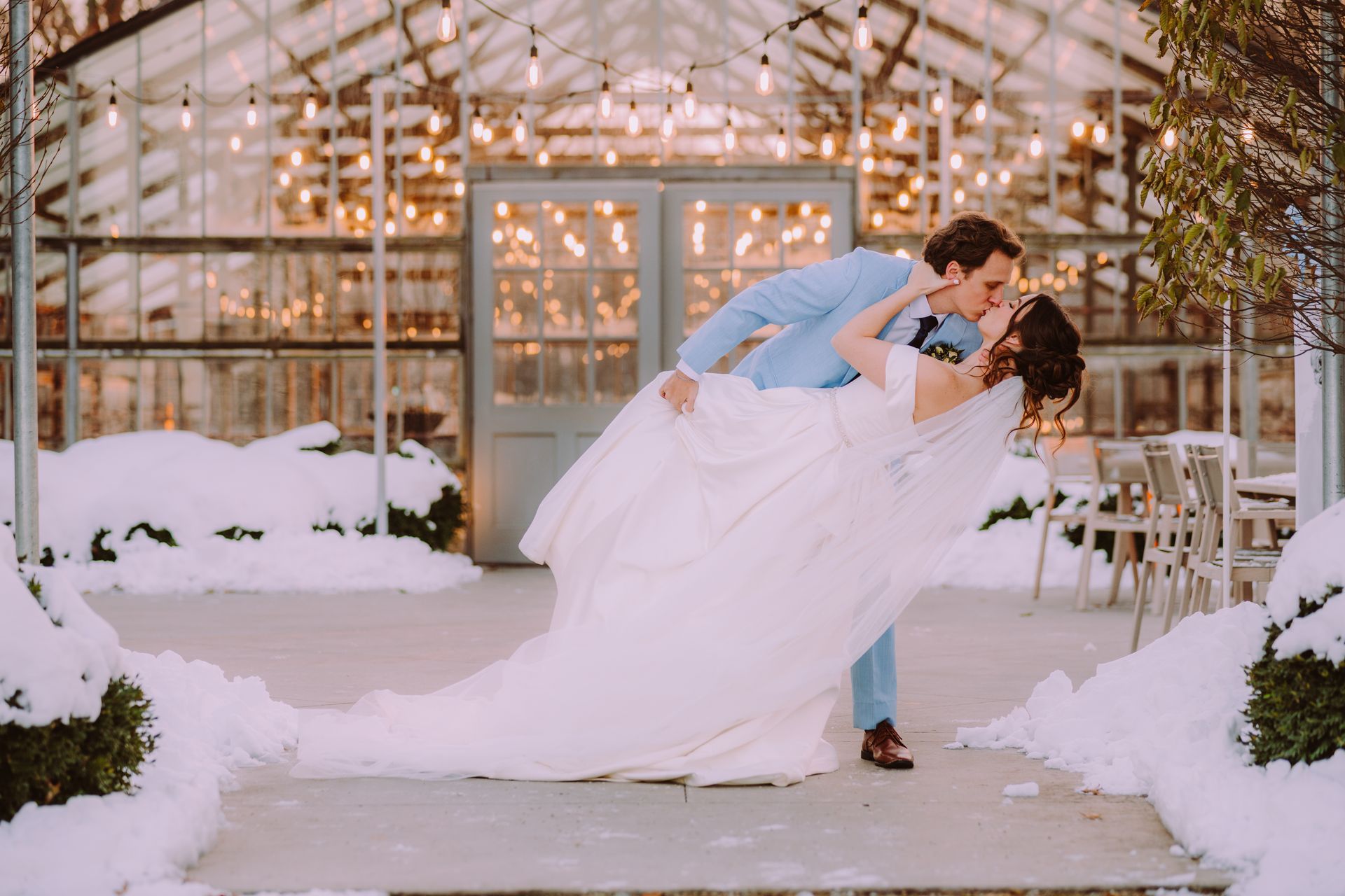 A couple kisses during a winter wedding. The groom dips the bride, snow covers the ground, lights twinkle in background.