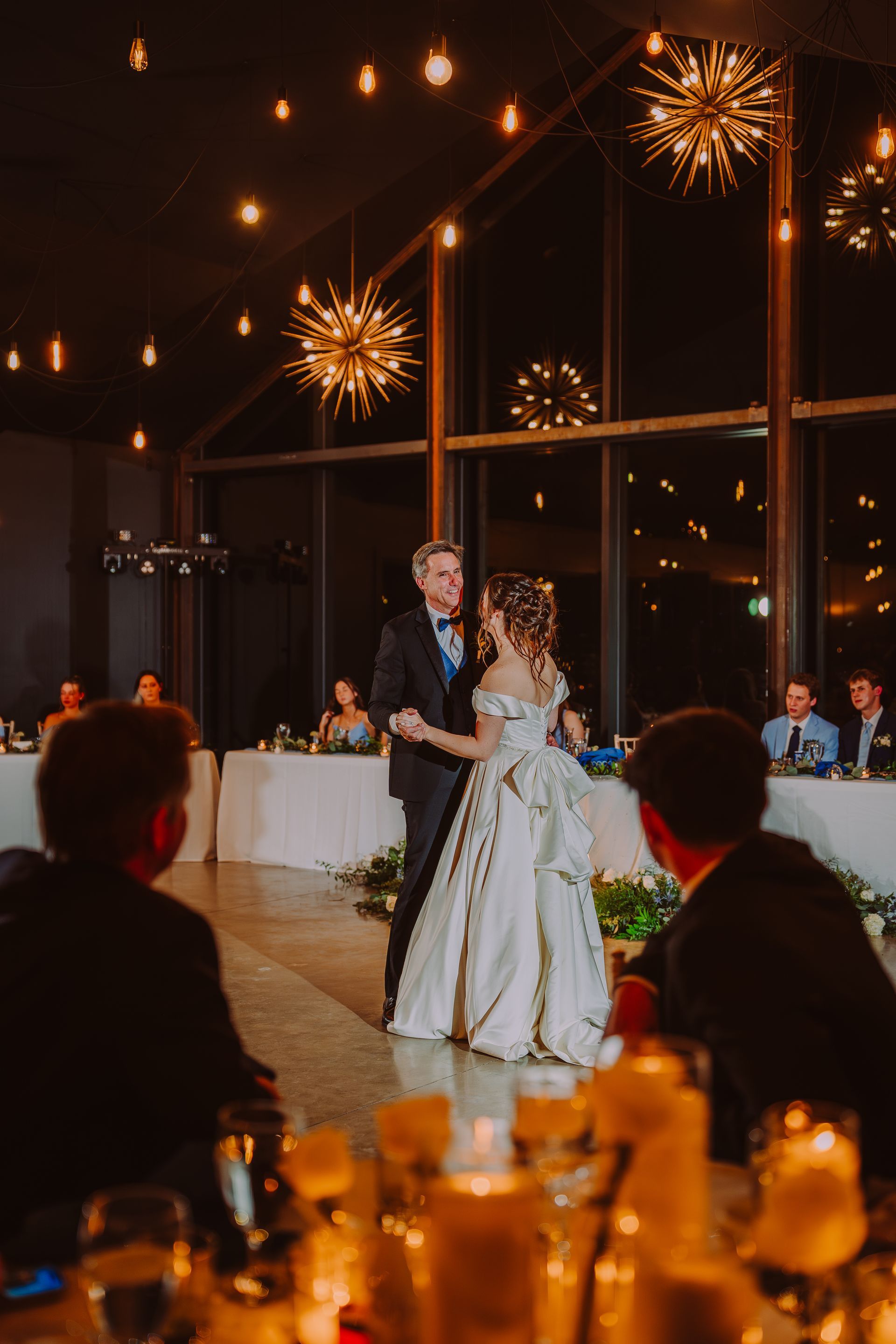 Bride and groom dance at reception. They face each other, lit by warm lights and surrounded by guests.