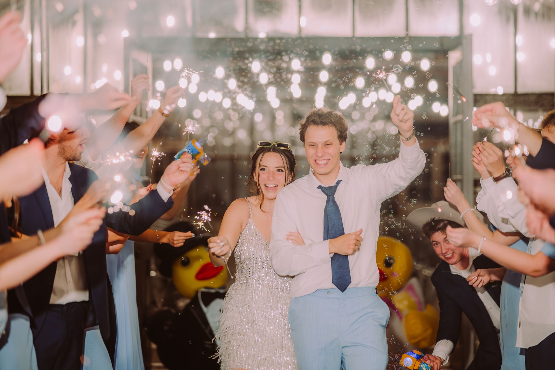 Couple celebrates with sparklers, arms raised, surrounded by cheering guests.