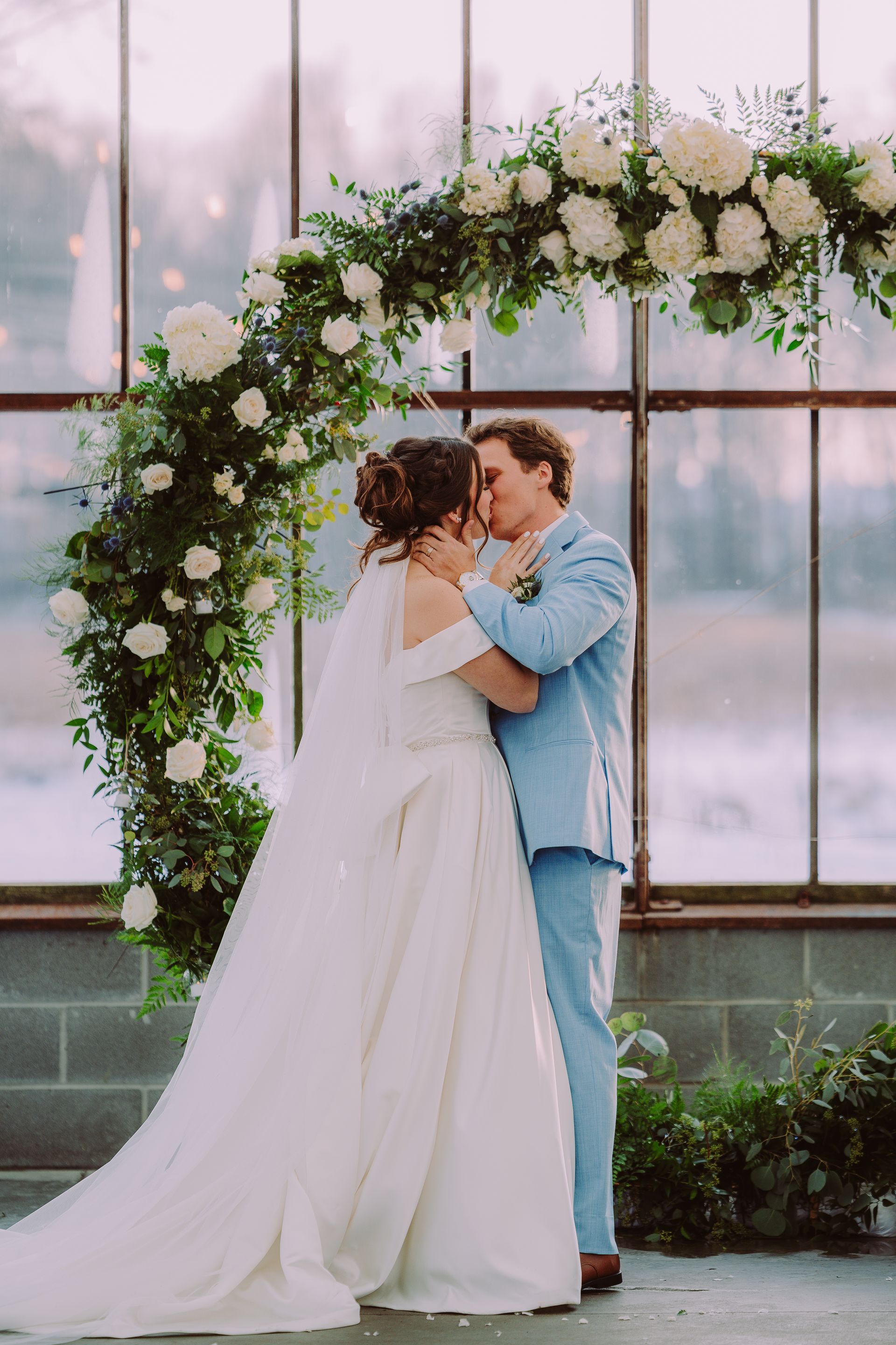Couple embraces under floral arch, wedding ceremony. Bride in white dress, groom in blue suit.