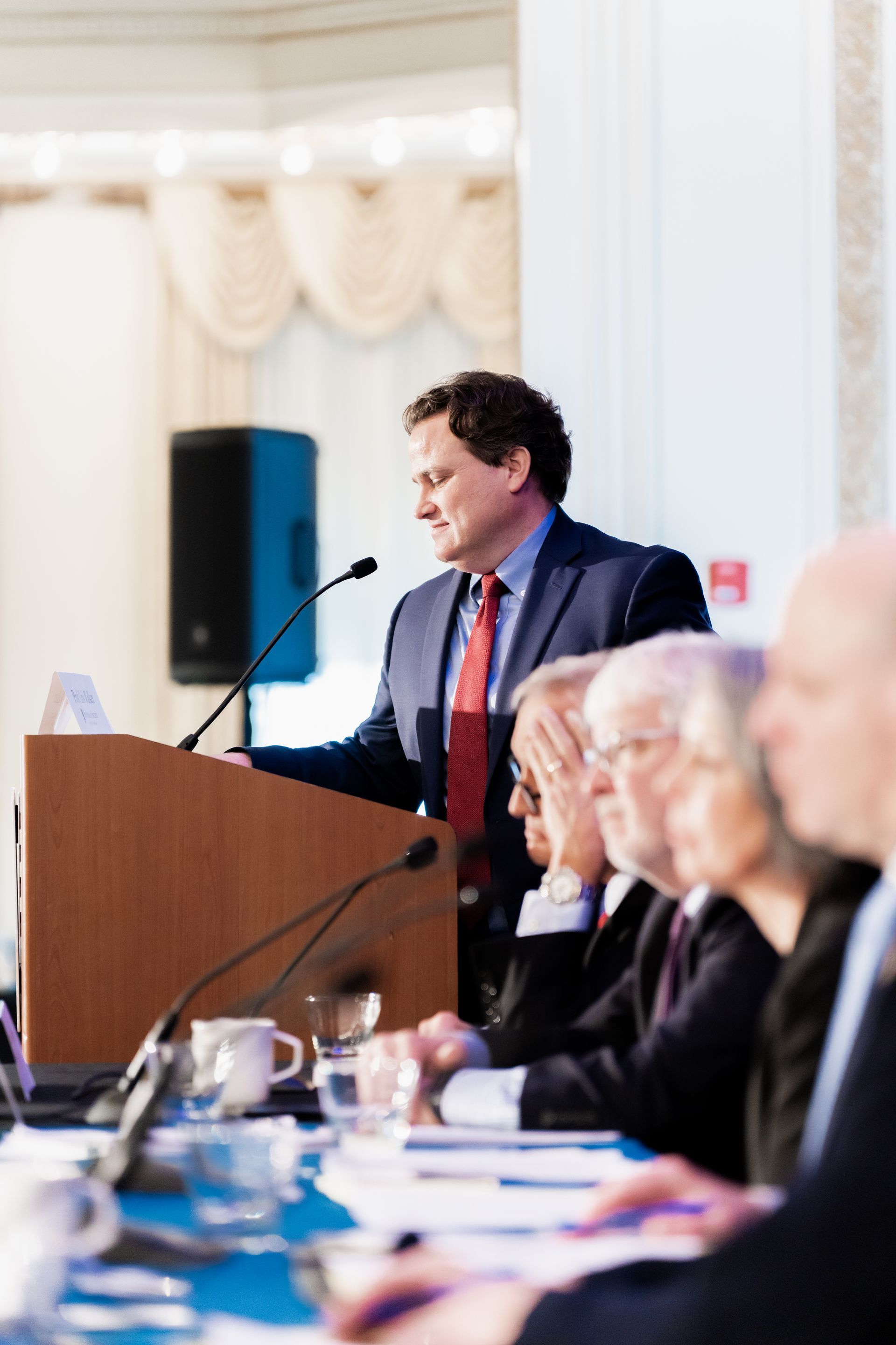 A man in a suit and red tie stands at a wooden podium speaking into a microphone before an audience at a table.
