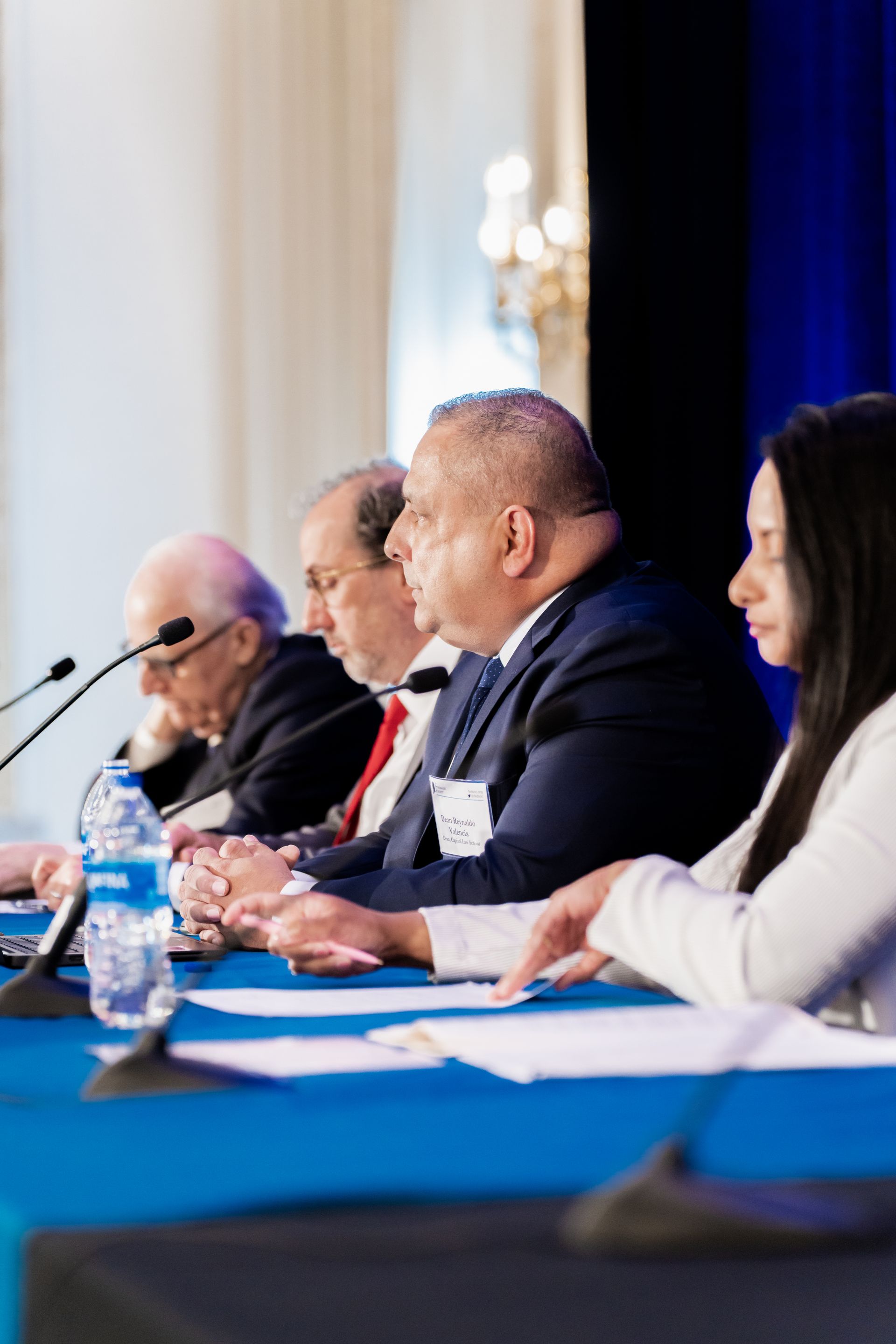 Four people sit at a long table with microphones during a conference, with a blue backdrop and papers in front of them.