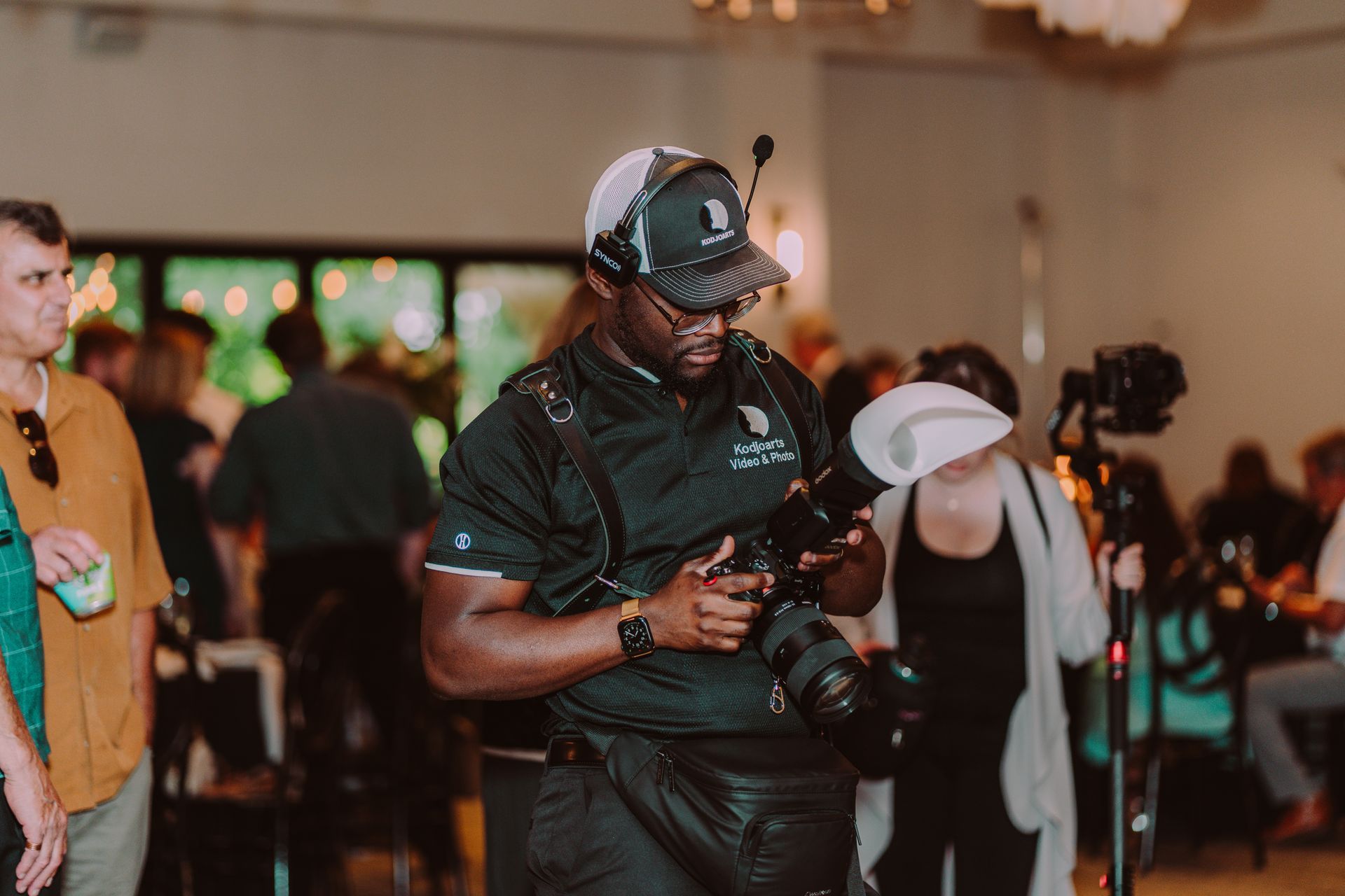 Photographer in a black uniform with a camera, at an event. He is focused on the camera. Guests in the background.