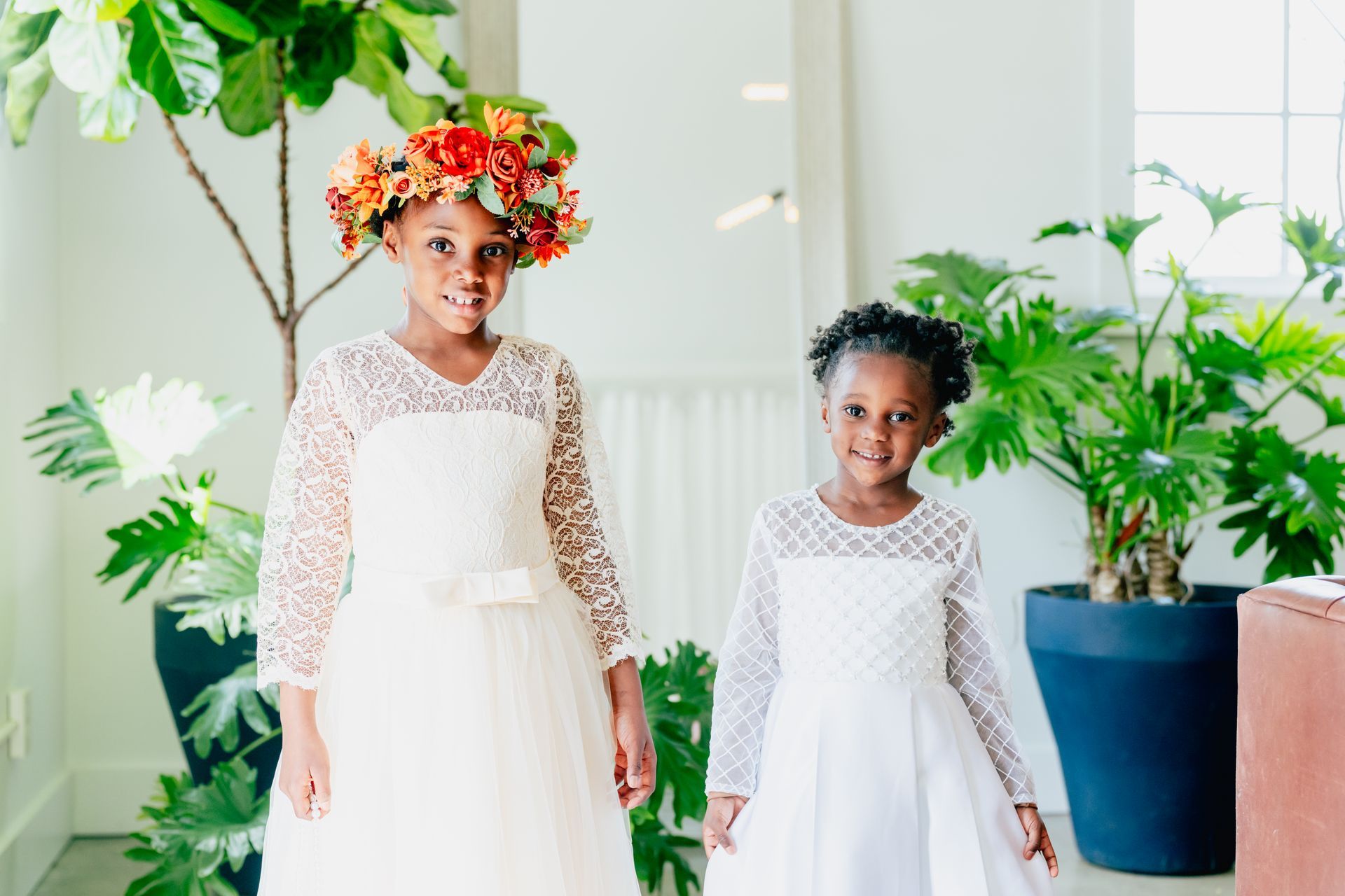 Two flower girls wearing white dresses and flower crowns are standing next to each other.
