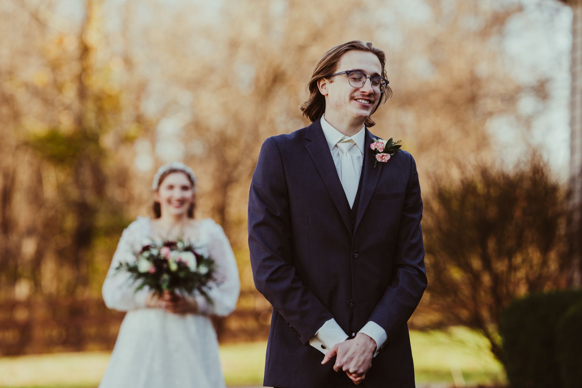 Groom smiles as bride approaches holding bouquet in outdoor setting.