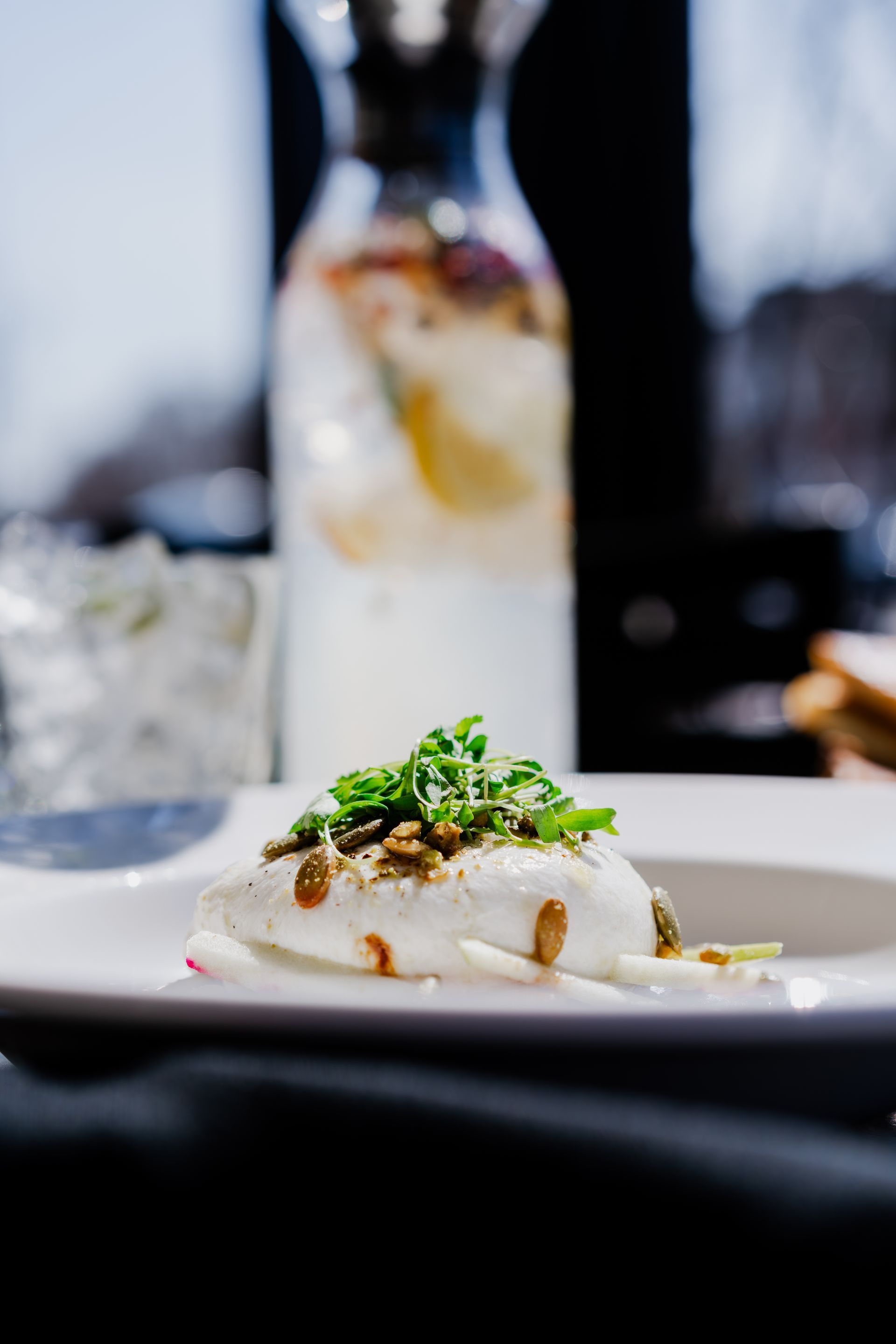 White dish with a creamy, white dish topped with greens and a drizzle, with a water pitcher in the background.
