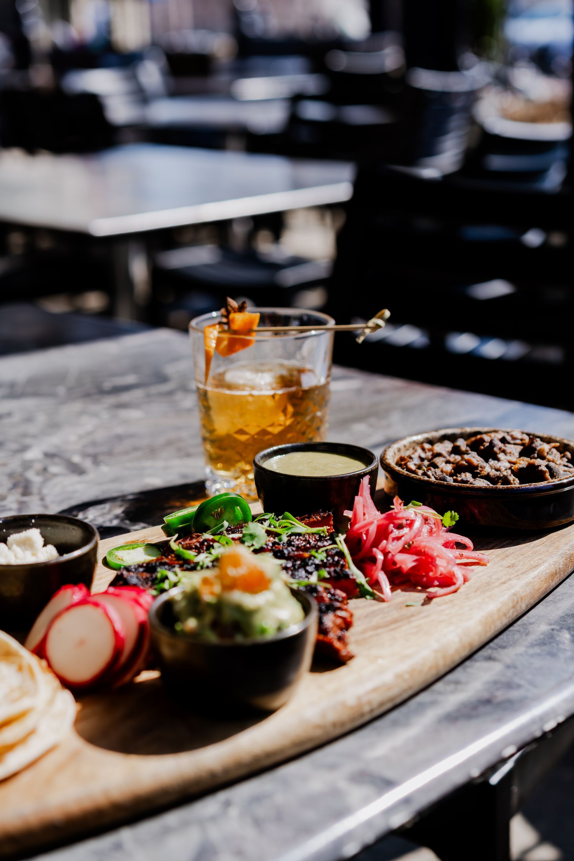Wooden board with tacos, small bowls of toppings, and an old-fashioned cocktail on a marble-topped table.