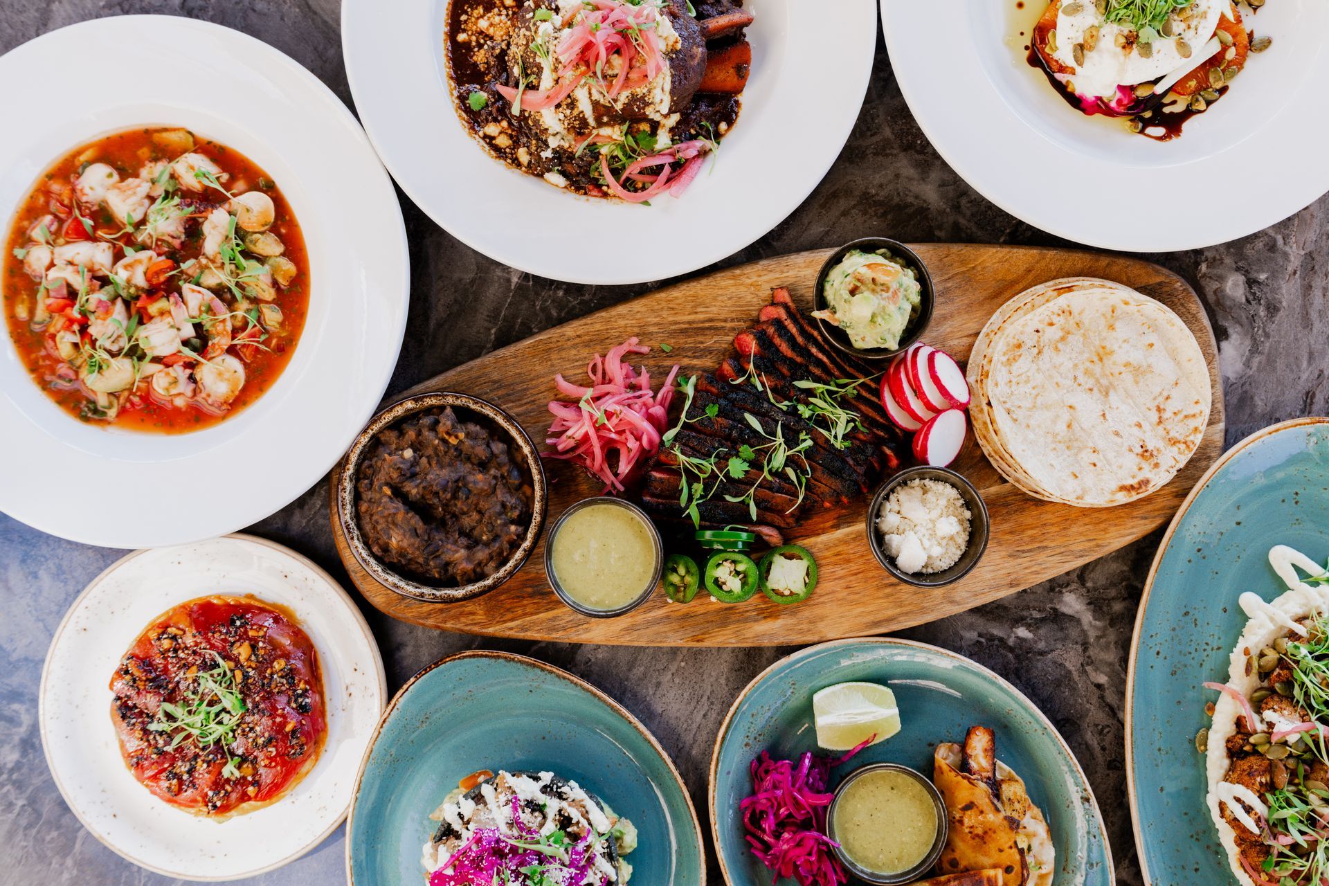 Assortment of Mexican dishes: ceviche, tacos, grilled meat, and sides on a table.