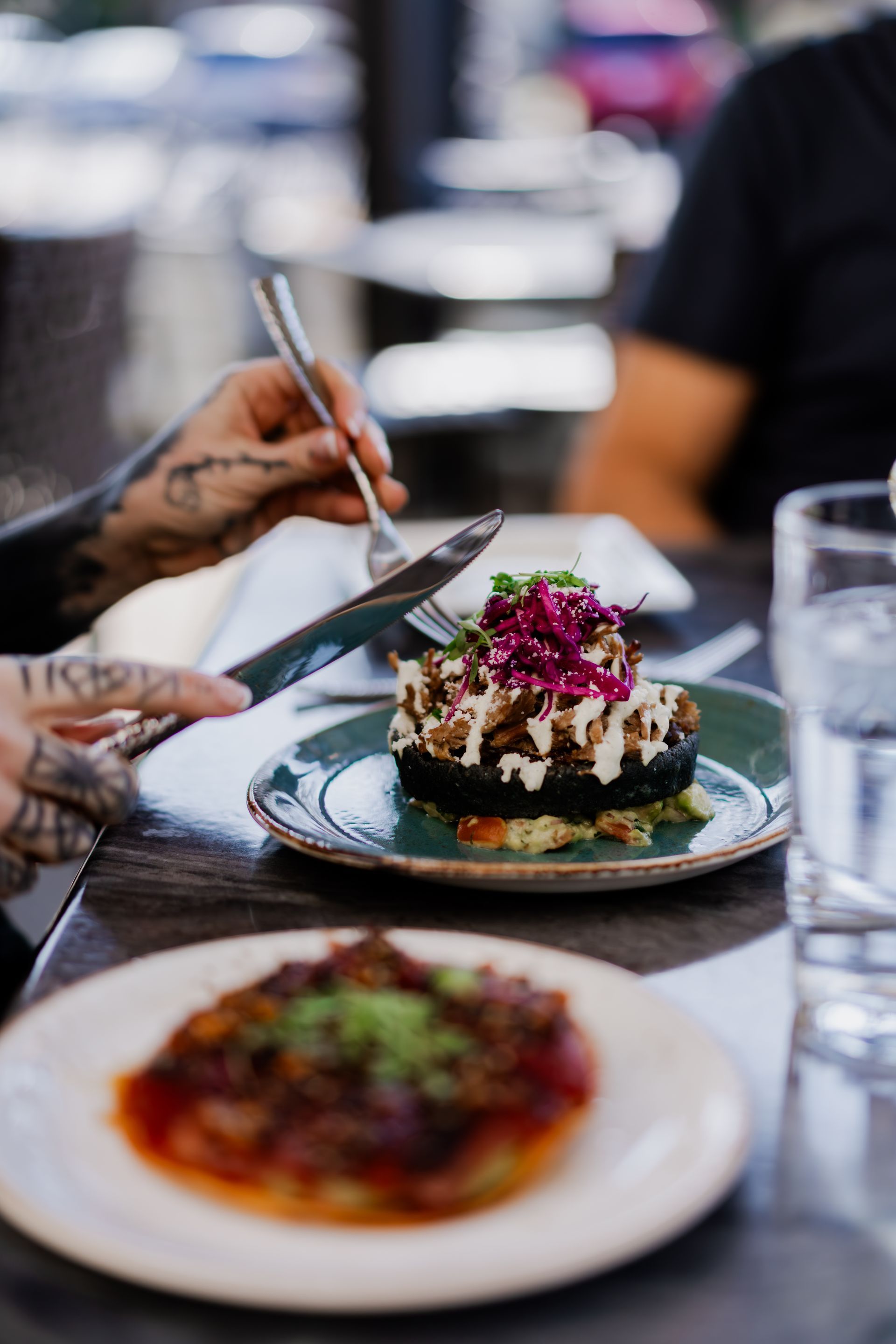 Person with tattoos eating a plate of food, outdoor dining, colorful dish with sauces and topping.