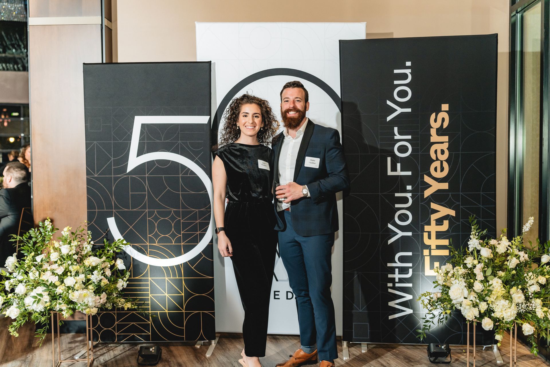 A man and a woman are standing in front of a sign that says `` with you for fifty years ''.