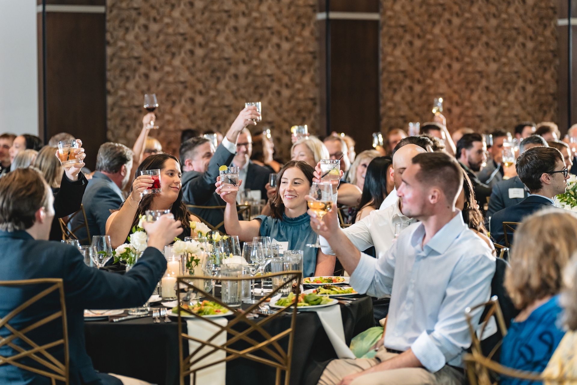 A large group of people are sitting at tables raising their glasses in the air.