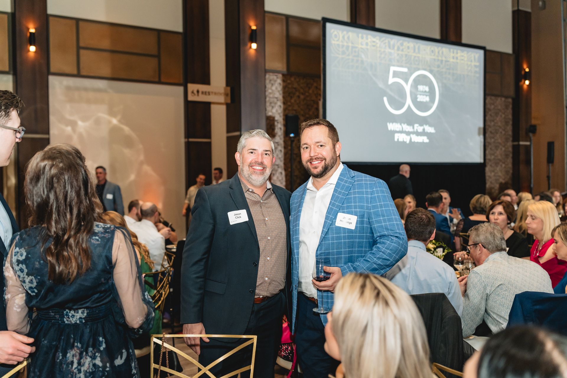 Two men are standing next to each other in front of a large screen at a party.