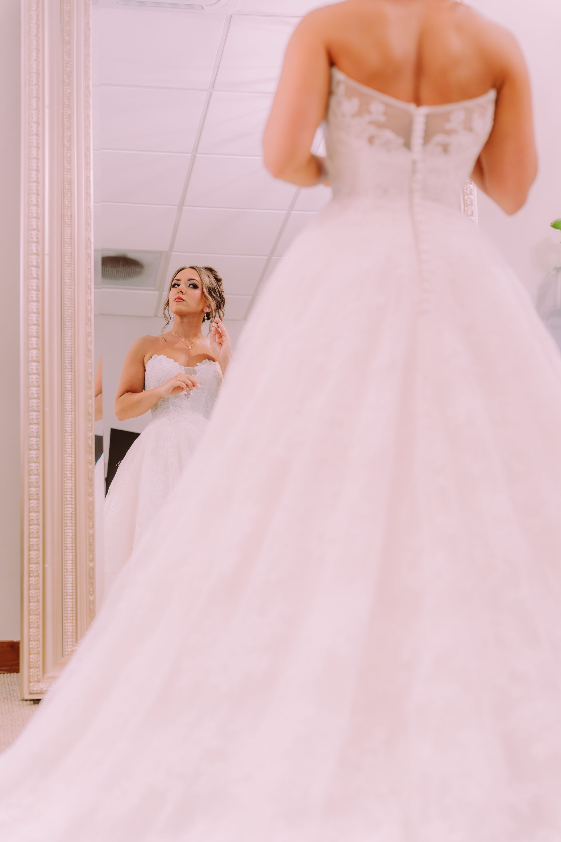Bride in a wedding dress, facing a mirror. She's adjusting her necklace in a bridal shop.