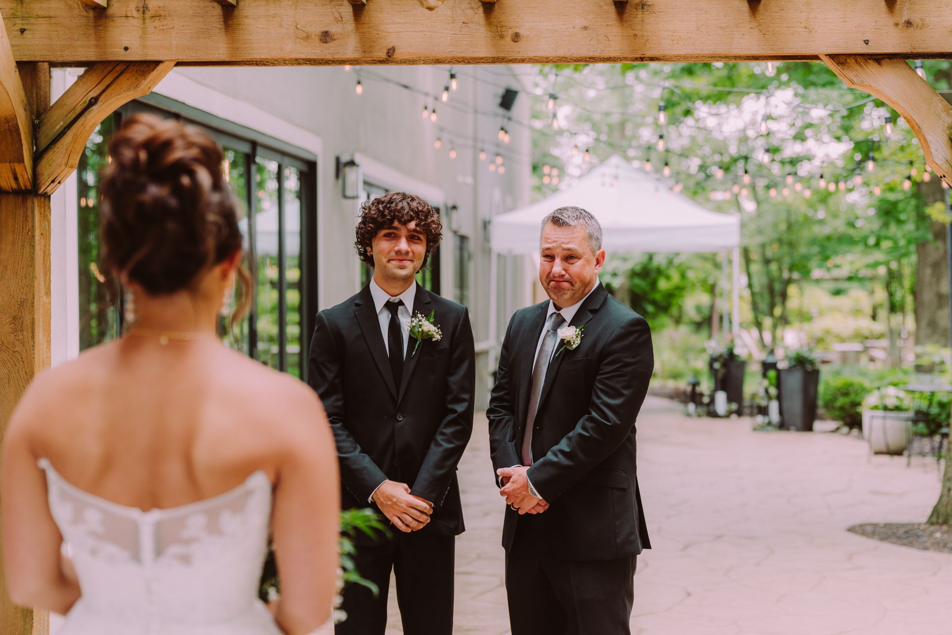 Bride facing groom and father, first look moment under an arbor. All three are formally dressed, the setting is outdoors.