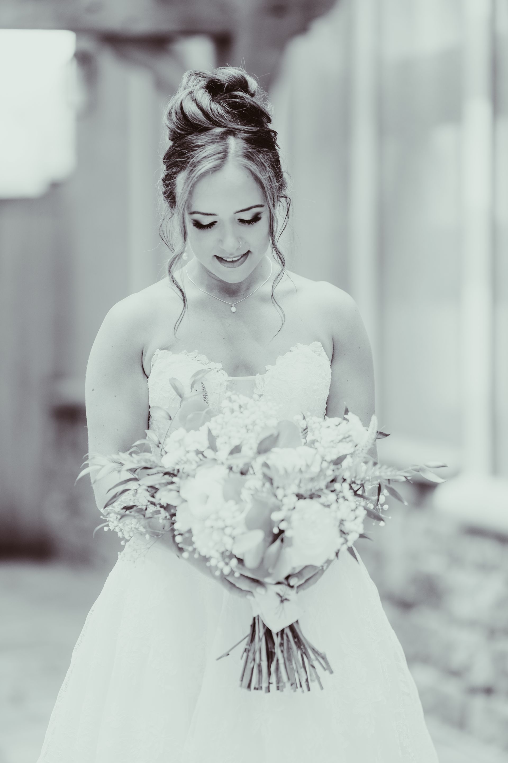 Bride in a strapless gown, holding a bouquet, looking down with a gentle smile. Black and white photo, outdoor setting.