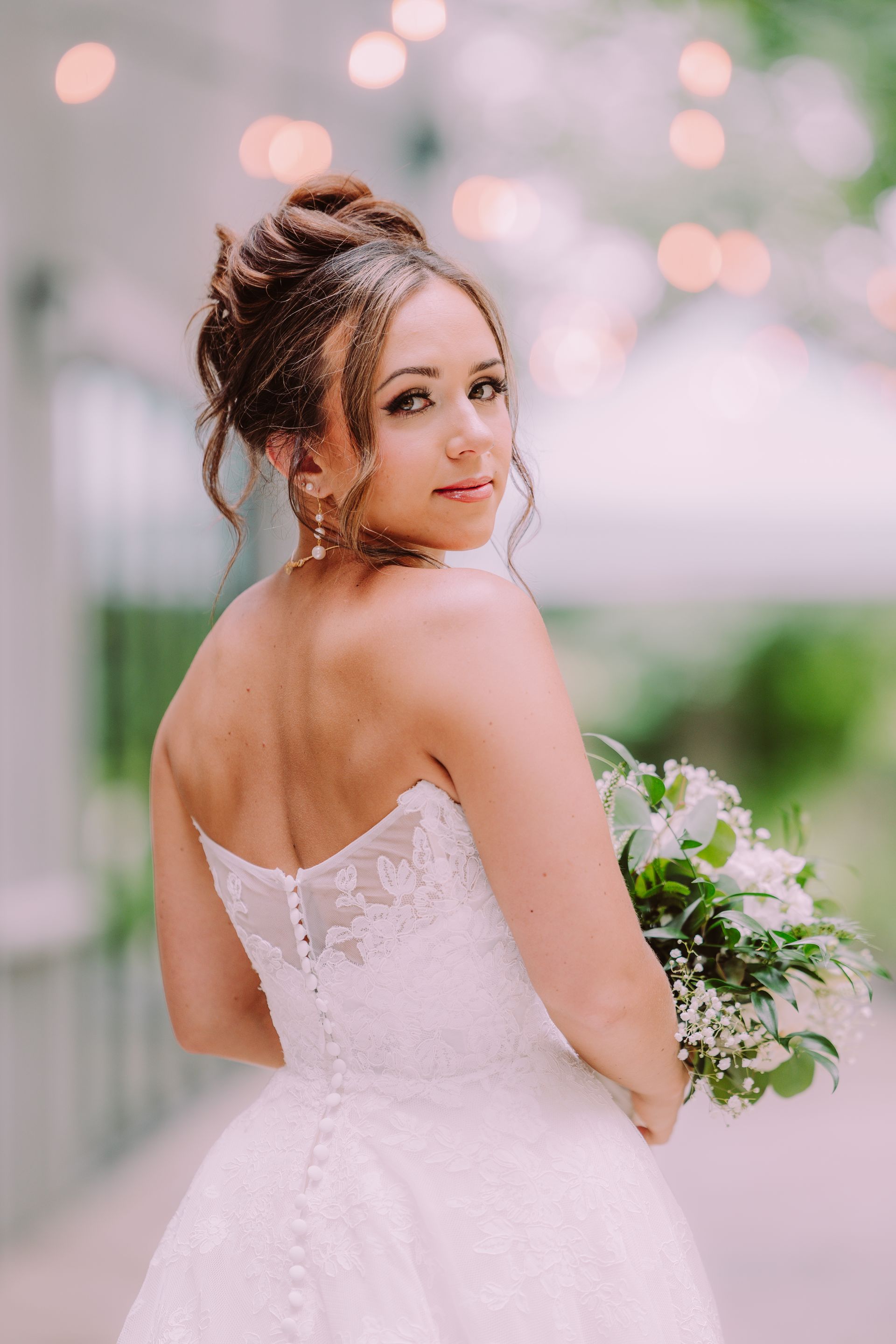 Bride in a white strapless gown, looking over her shoulder, holding a bouquet; outdoors with string lights in the background.