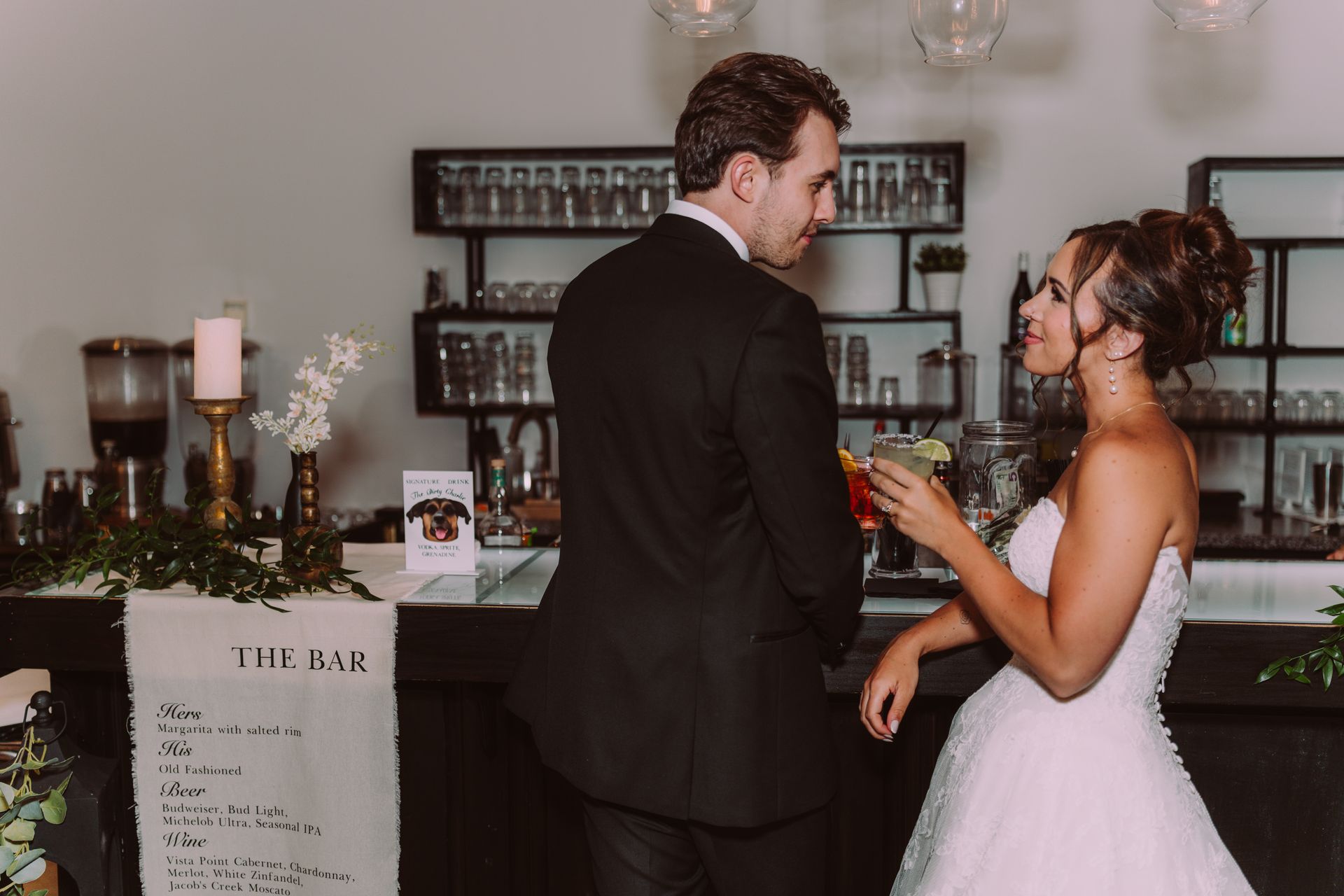 Bride and groom at a bar, she holds a cocktail, smiling at him. He looks back with a serious expression. Modern bar setting.