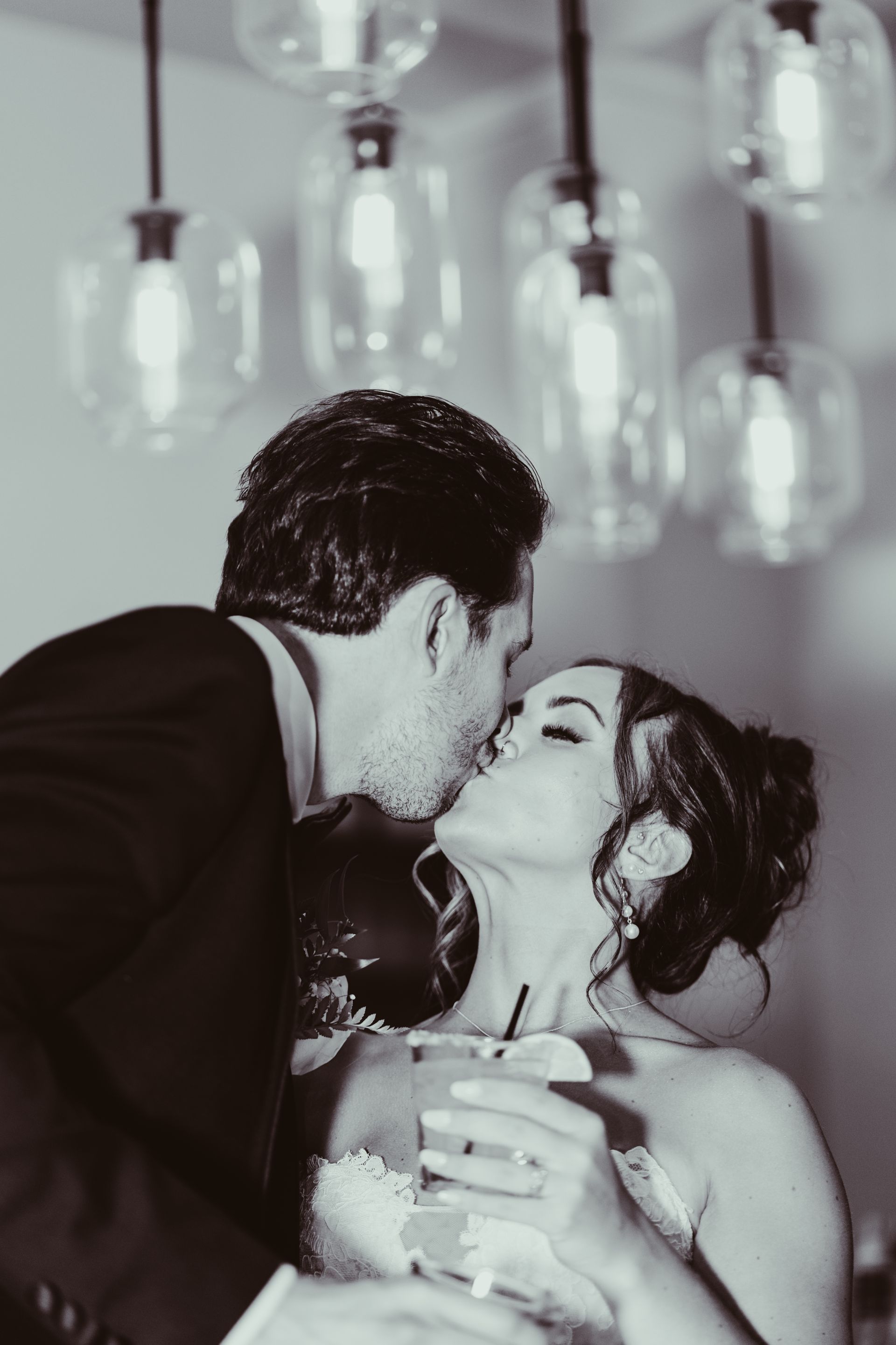 Bride and groom sharing a kiss, holding a cocktail in a bar with hanging lights. Black and white.