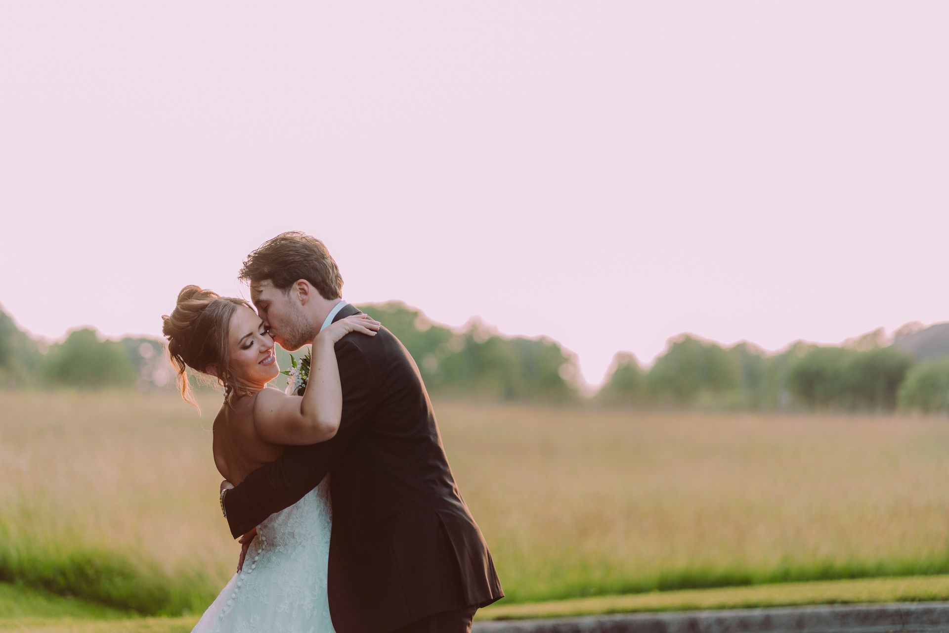 Newlyweds embrace outdoors at sunset. The bride in a white gown smiles, while the groom in a black suit kisses her forehead.