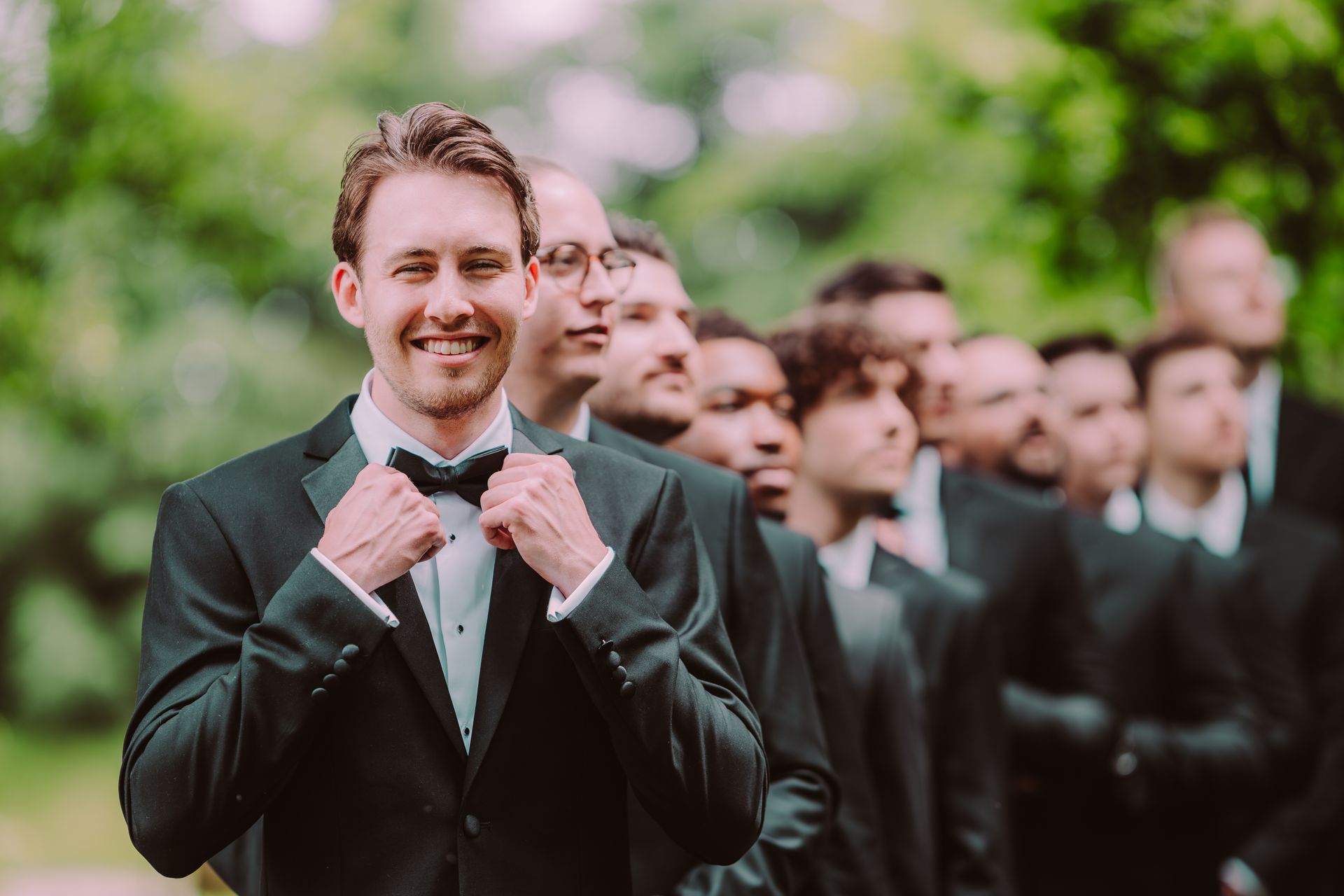 Groom in a black tuxedo smiles, adjusting his bow tie, with groomsmen lined up behind him in a lush green outdoor setting.