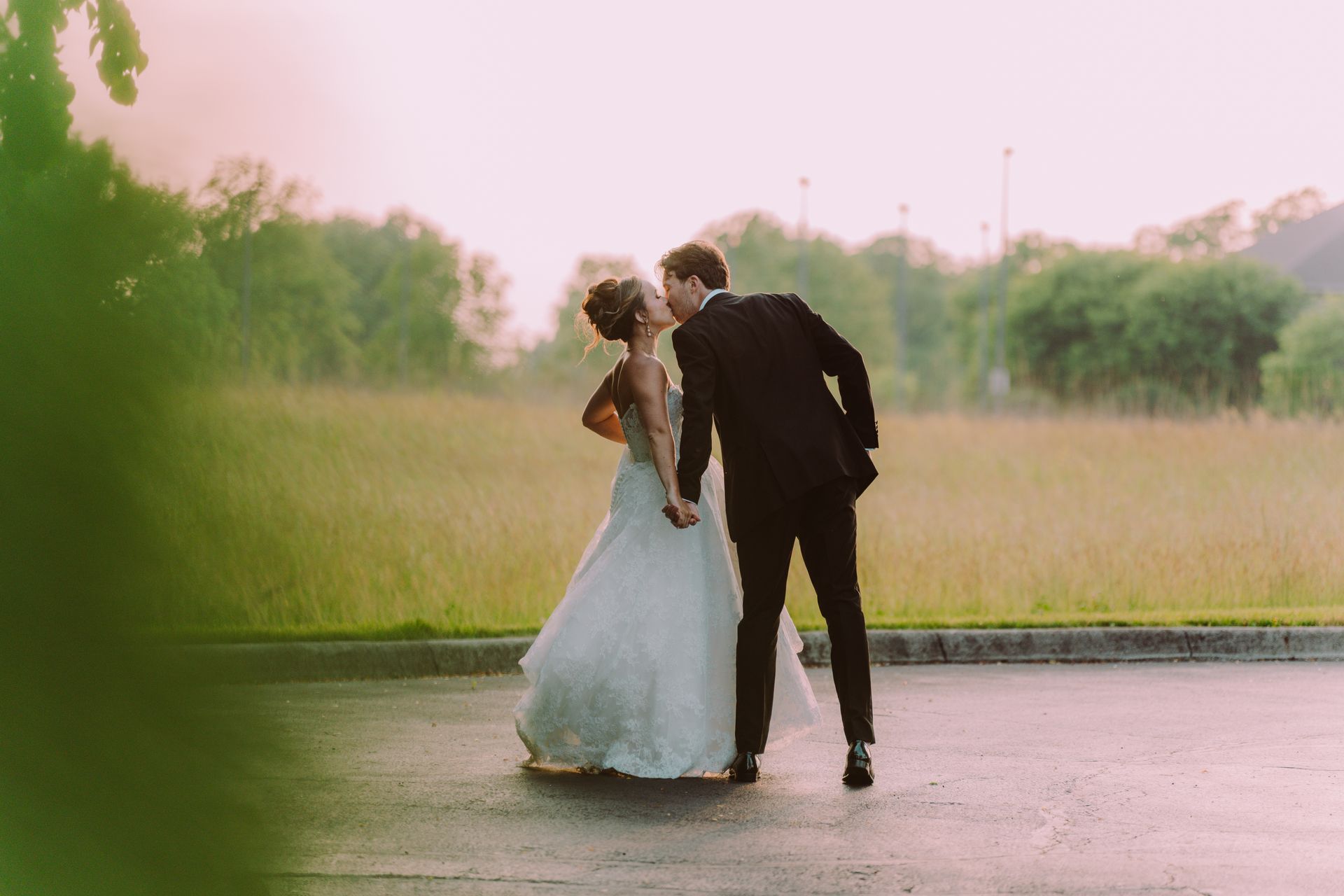A newlywed couple kissing outdoors at sunset. The bride wears a flowing, light blue dress, and the groom wears a black suit.