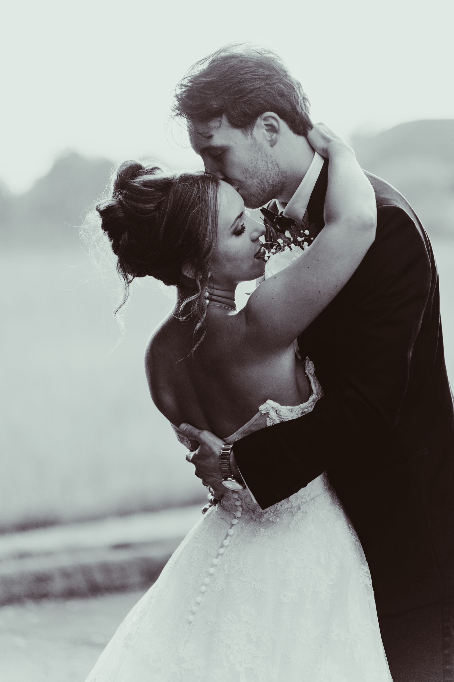 Bride and groom embracing, groom kissing bride's forehead. They are outdoors in a field, soft focus, black and white.