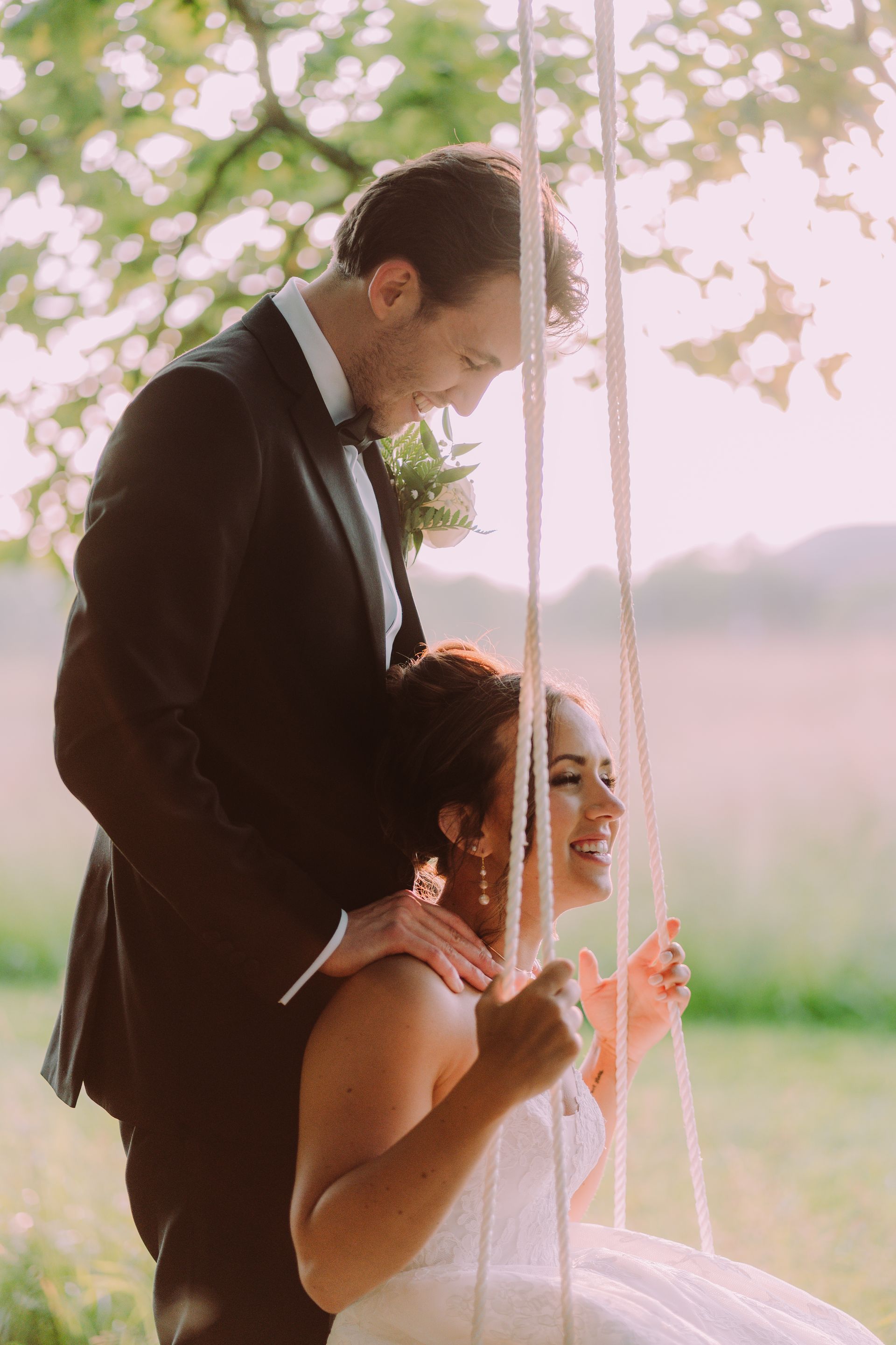 Newlywed couple on a swing outdoors; the groom looks lovingly at the smiling bride. Soft light bathes the scene.