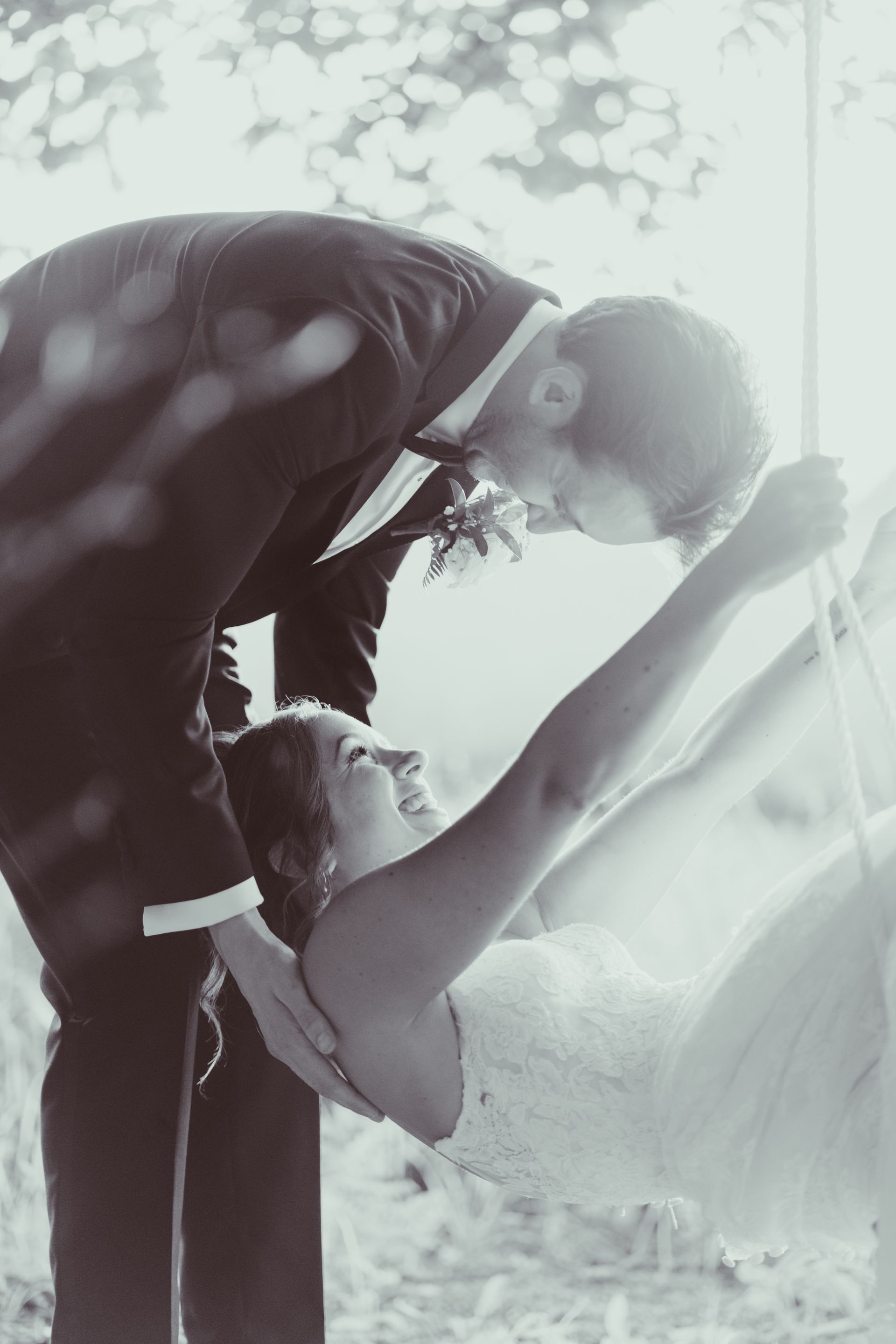 Groom leaning in to kiss bride on a swing. They are in formal wear outdoors, a grayscale image.