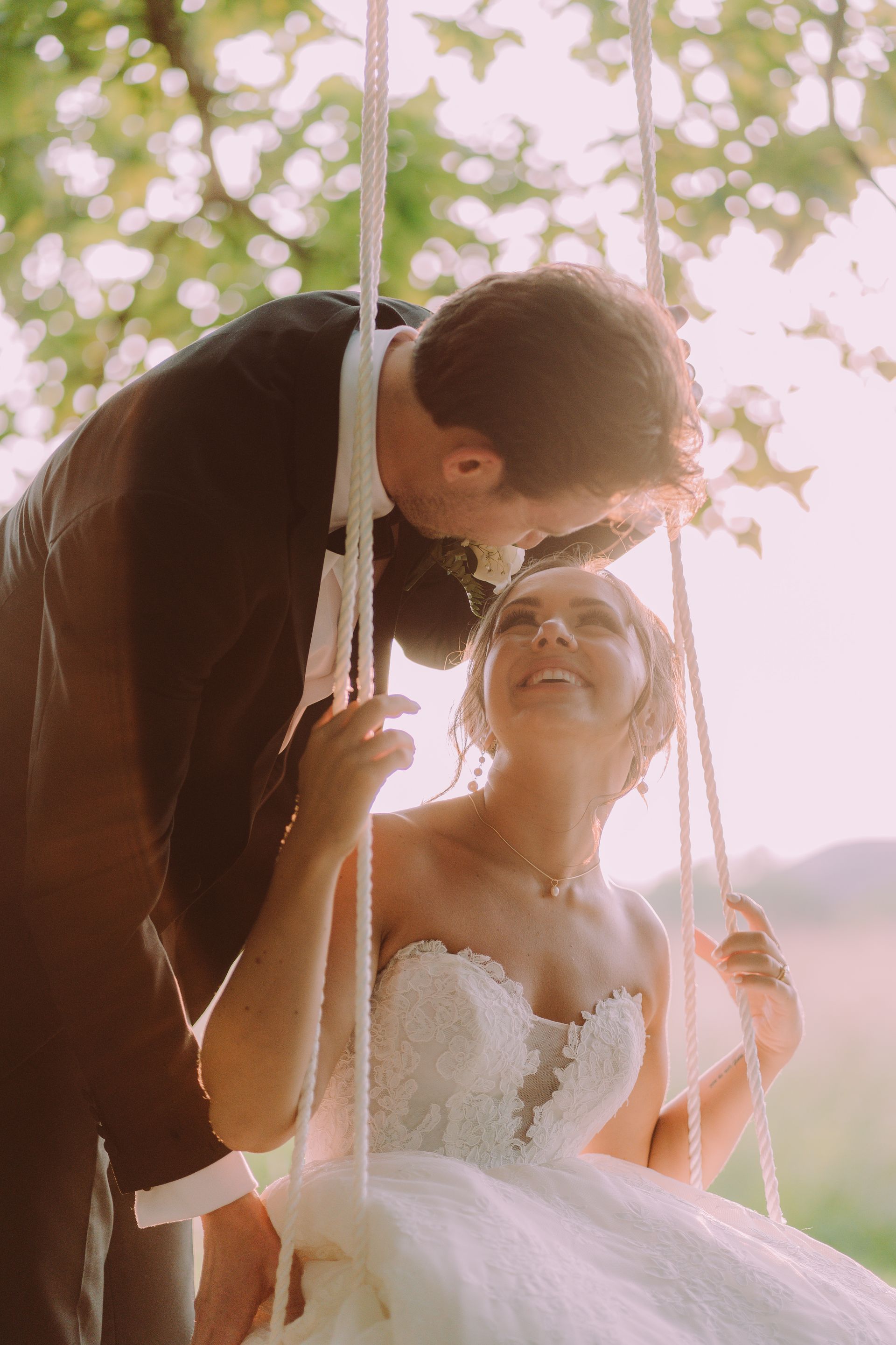 A bride and groom on a swing, smiling at each other. She wears a white dress; he wears a suit. Outdoors, with sunlight.