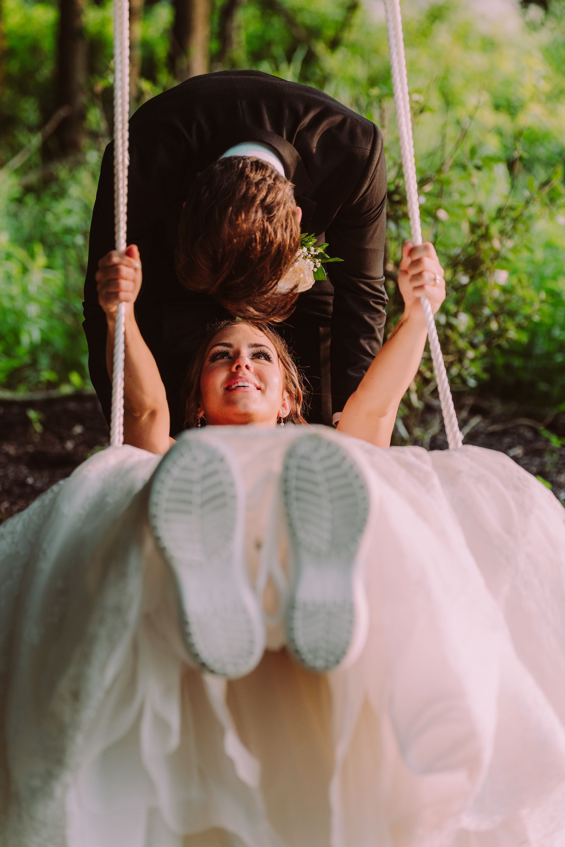 Bride on swing, wearing sneakers, groom pushing her. Forest backdrop, joyful expressions.