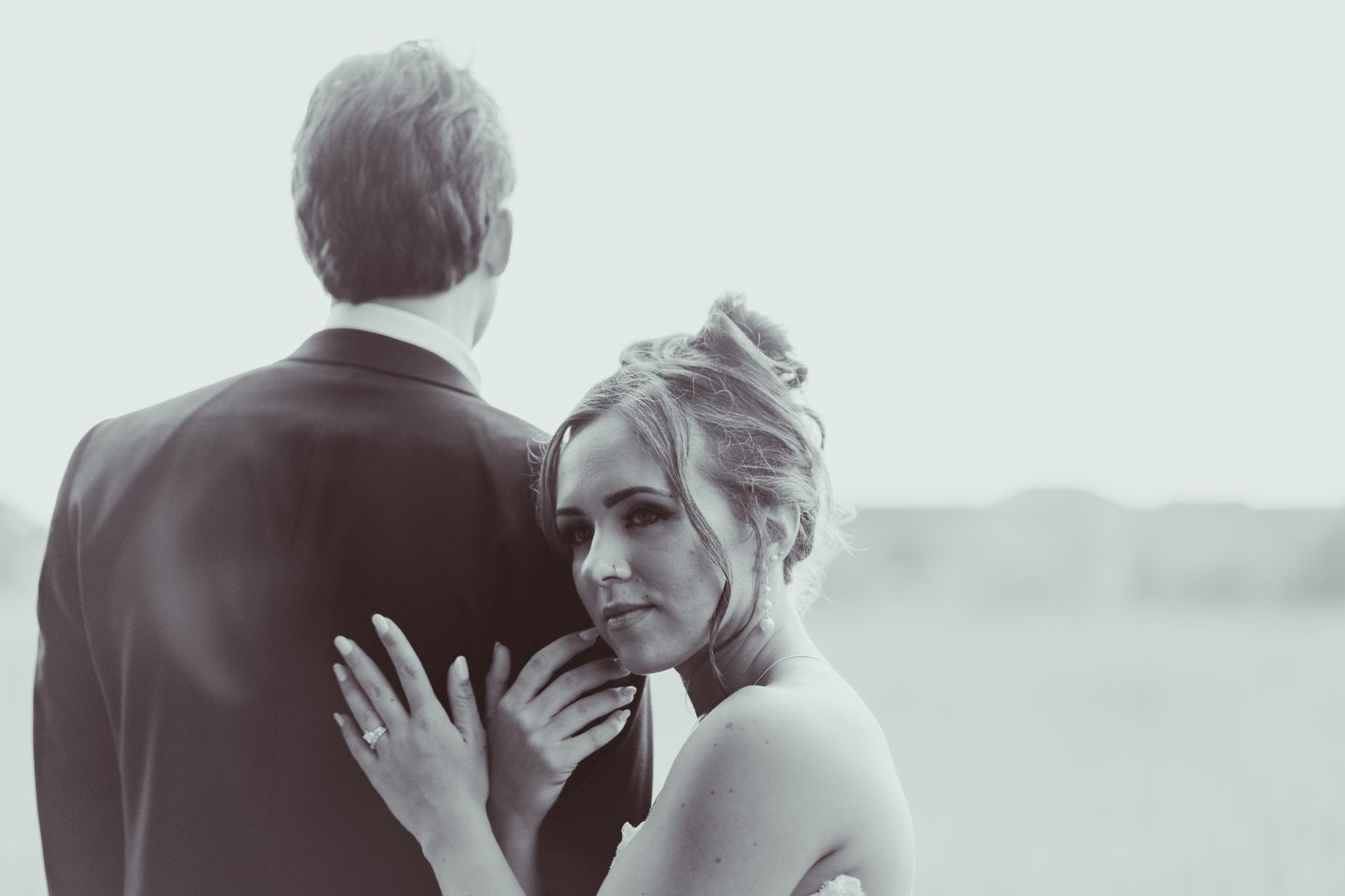 Bride in a wedding dress leans on the groom's back, looking at the camera with a tender expression; outdoors.