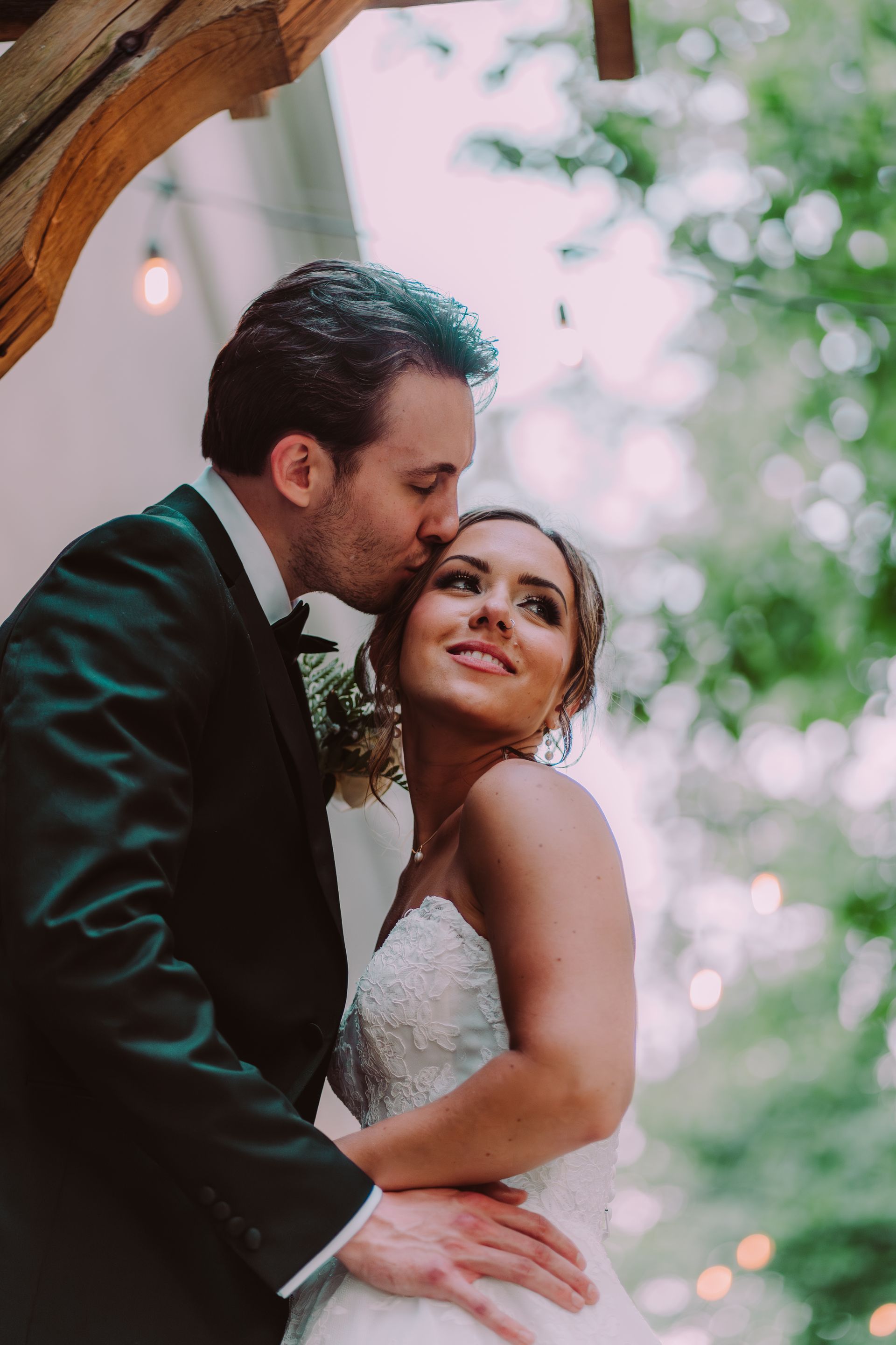 Groom kisses bride's forehead outdoors. She wears a white dress and smiles, looking up. He is in a tuxedo.