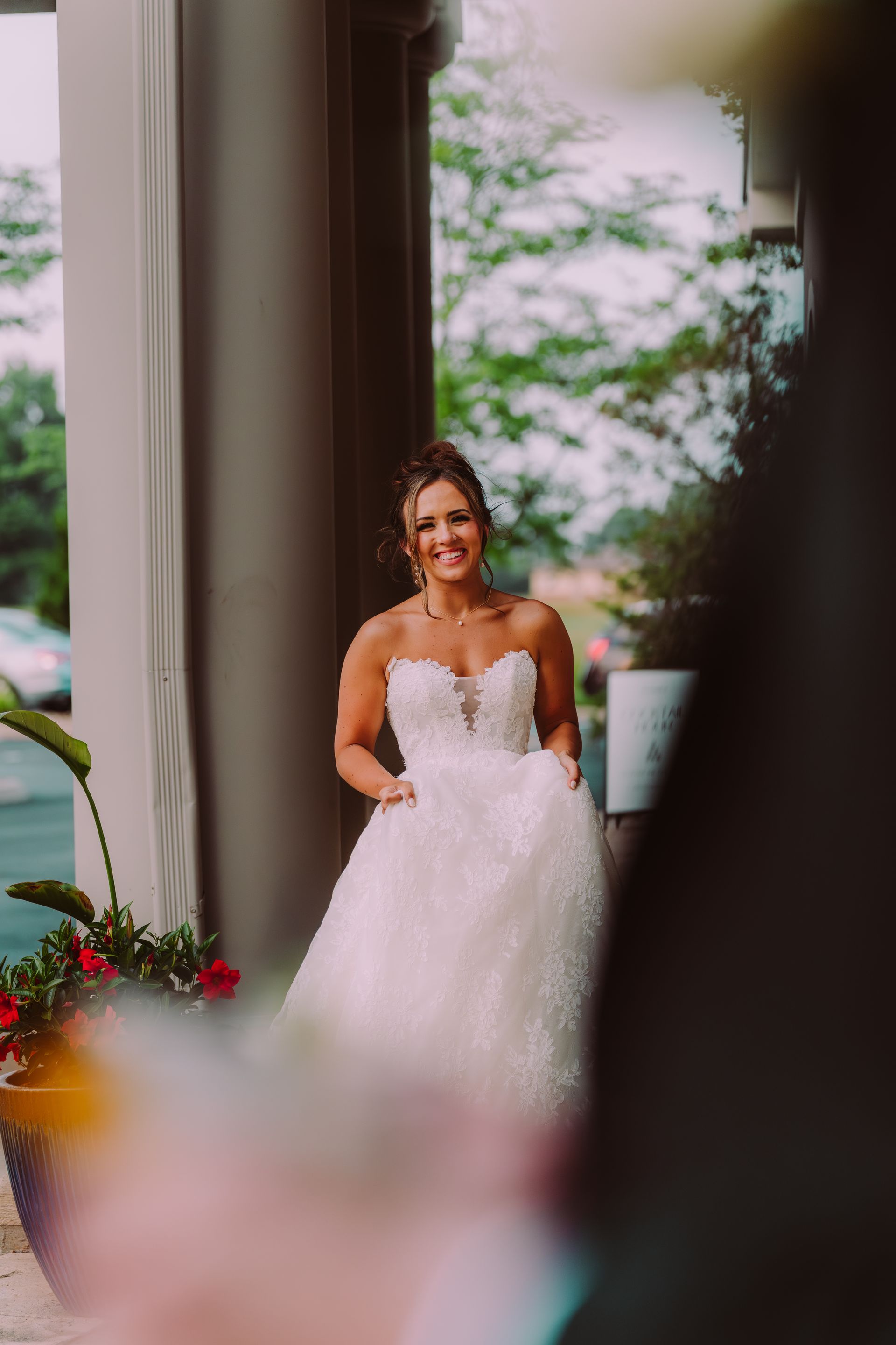 Bride in a white strapless gown smiles while posing outside, framed by a doorway and greenery.