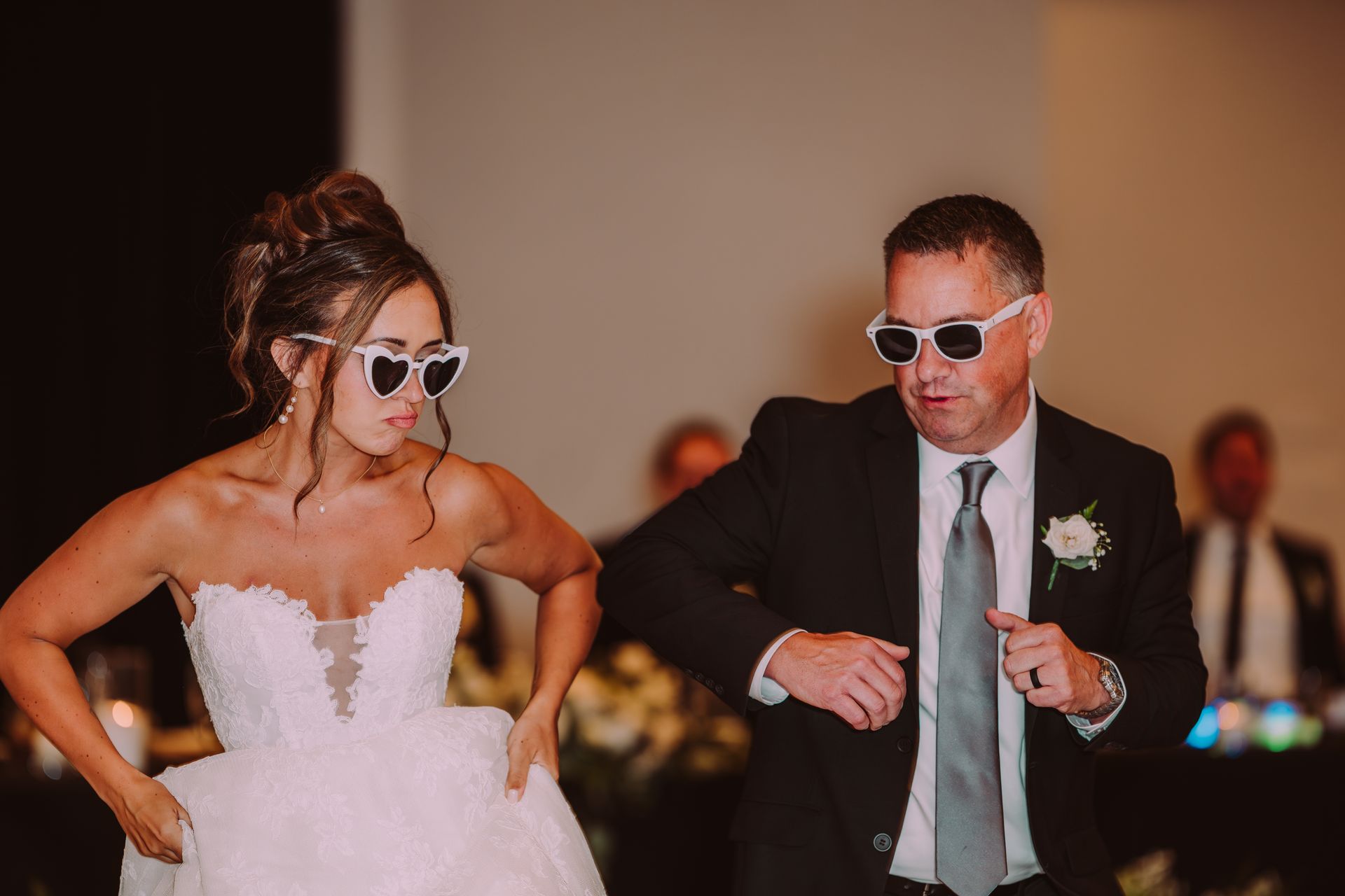 Bride and father dance at a wedding reception, both wearing white sunglasses. They are in a ballroom.